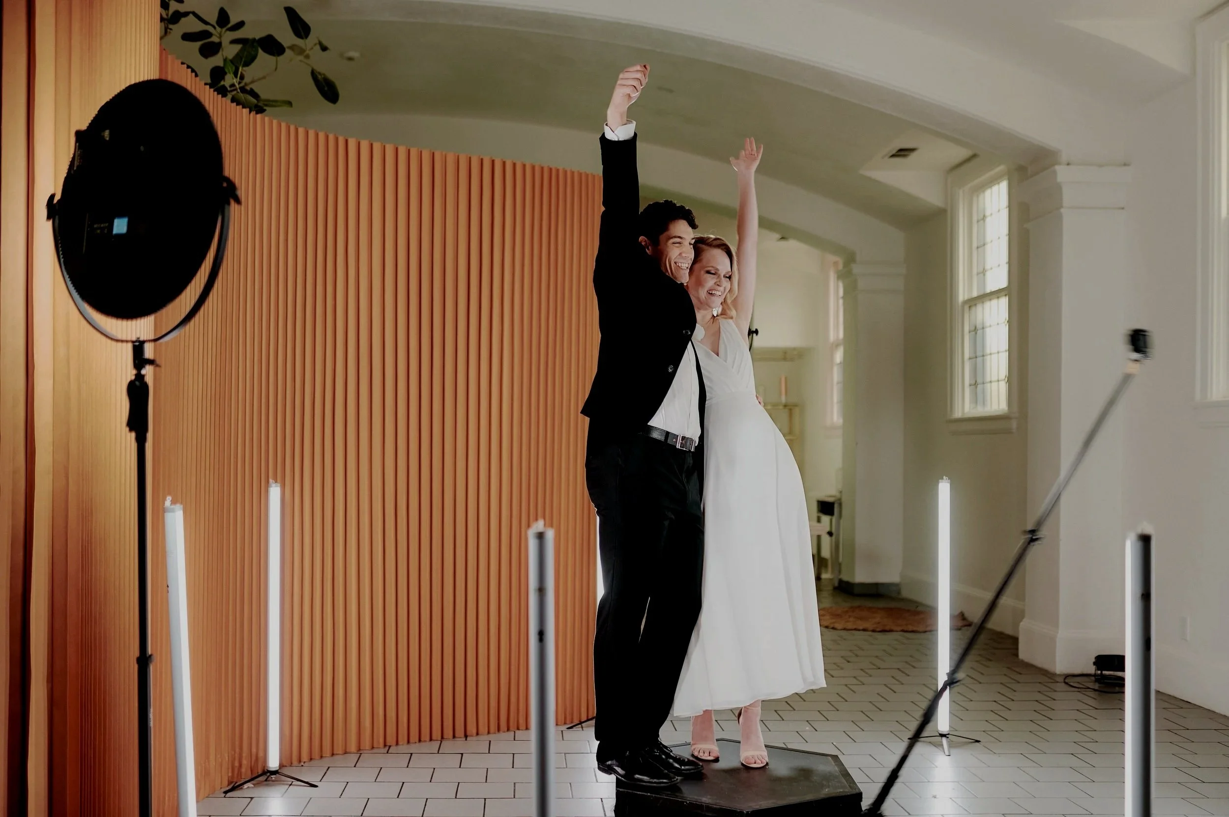 A well-dressed couple, a man in a black suit and a woman in a white dress, stand on a small platform with their arms raised in celebration inside a bright room with large windows. A photographer's studio light is visible to the left. 360 photo pop ph