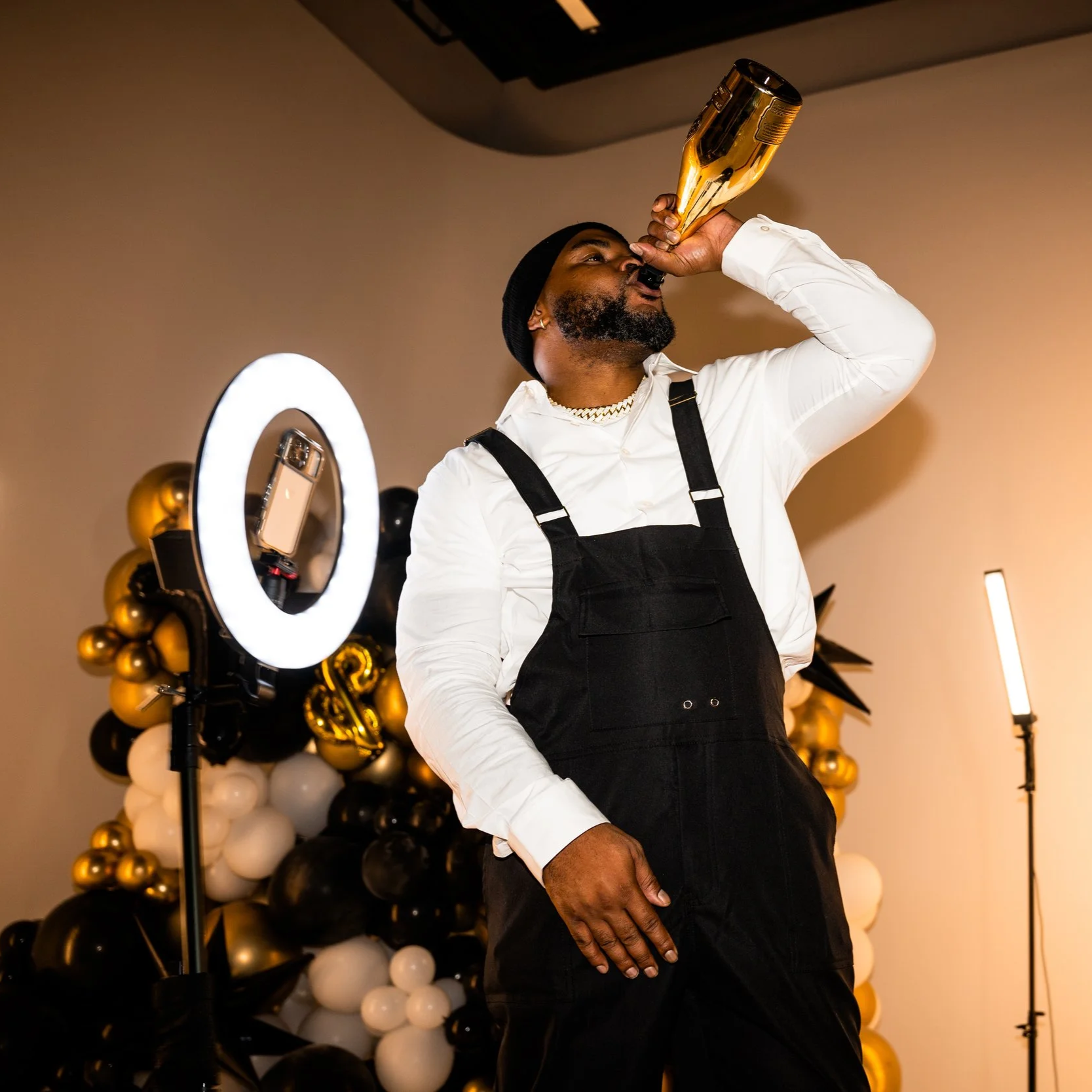A man wearing a black beanie and a white shirt with black overalls, holding a large gold bottle to his lips, standing in front of black, white, and gold balloons, with a ring light and a smartphone on a tripod nearby. 360 photo pop photo booth boise