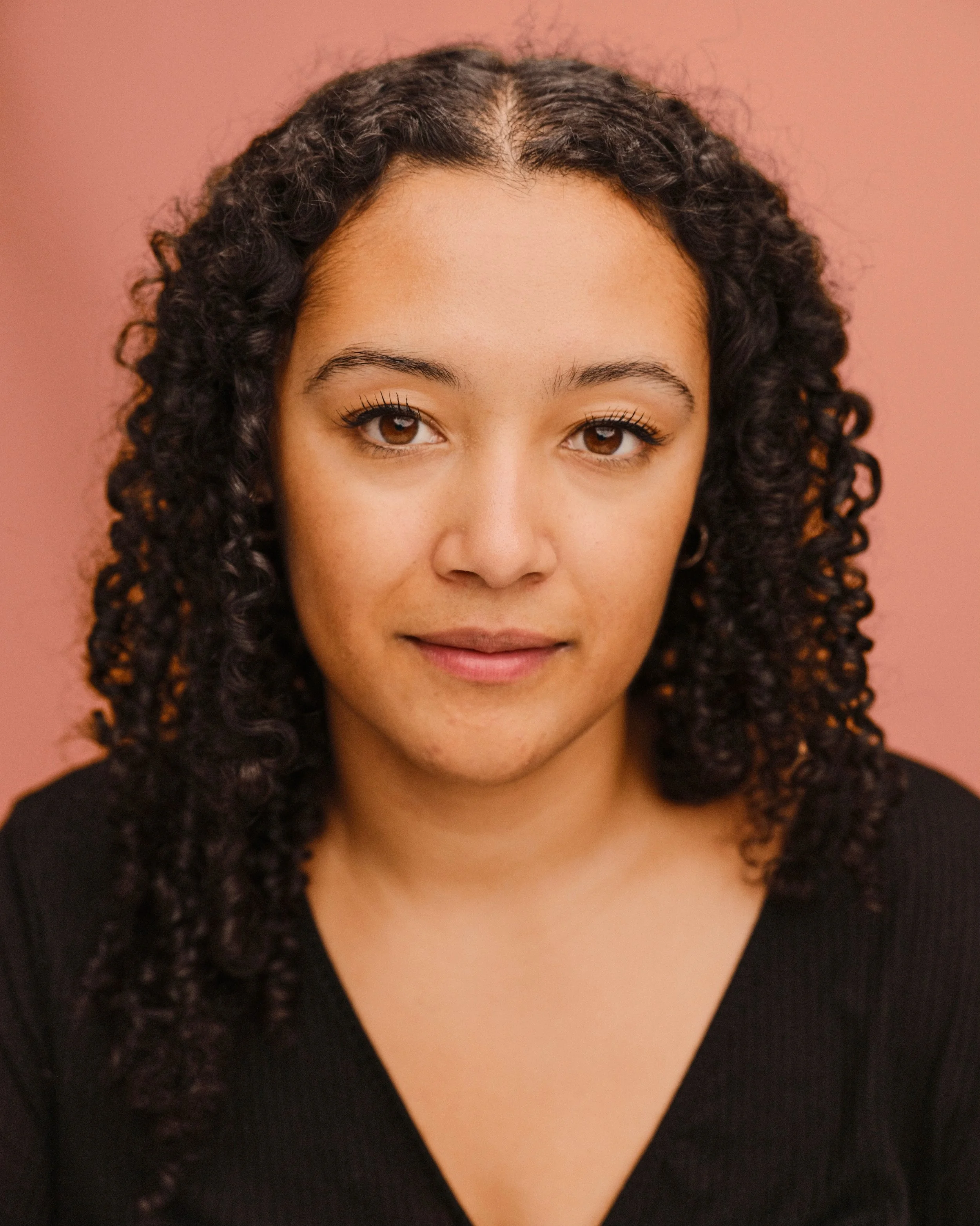 A young woman with curly hair smiling at the camera, wearing a black v-neck shirt.