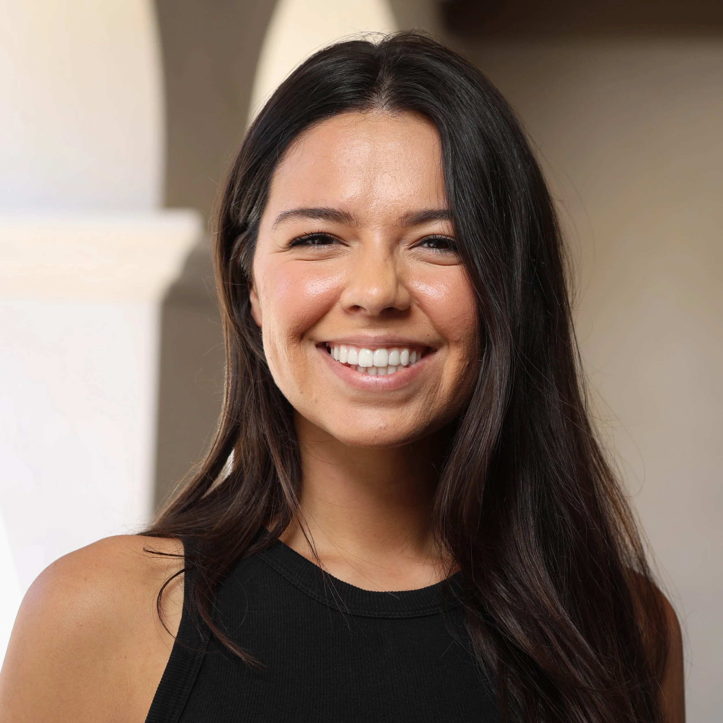 Smiling woman with long brown hair, wearing a navy blue shirt and hoop earrings, standing against a beige wall.