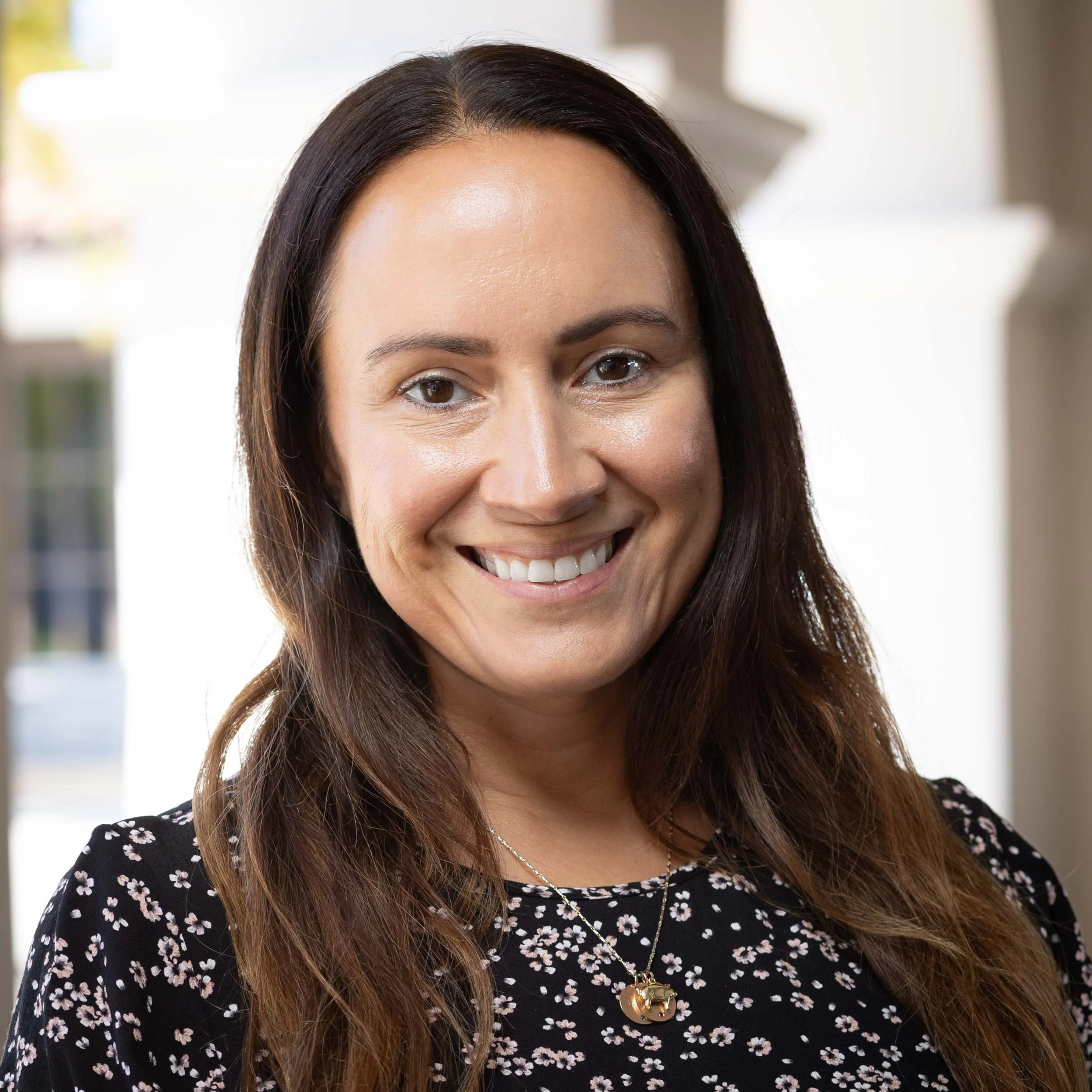 A woman with long brown hair wearing a navy blue polo shirt, smiling against a plain light-colored wall.