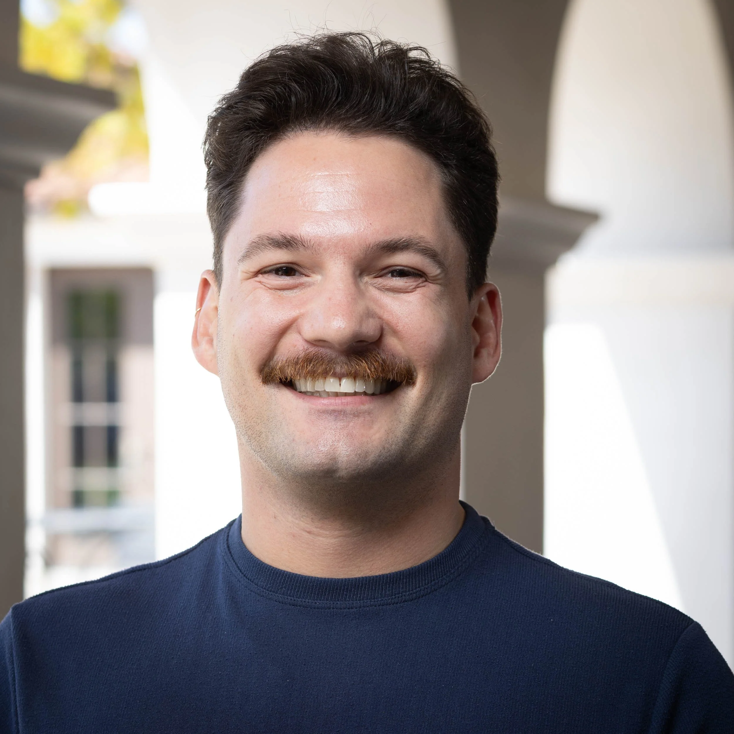 A man with dark hair, a mustache, and a blue collared shirt standing against a plain light-colored background.