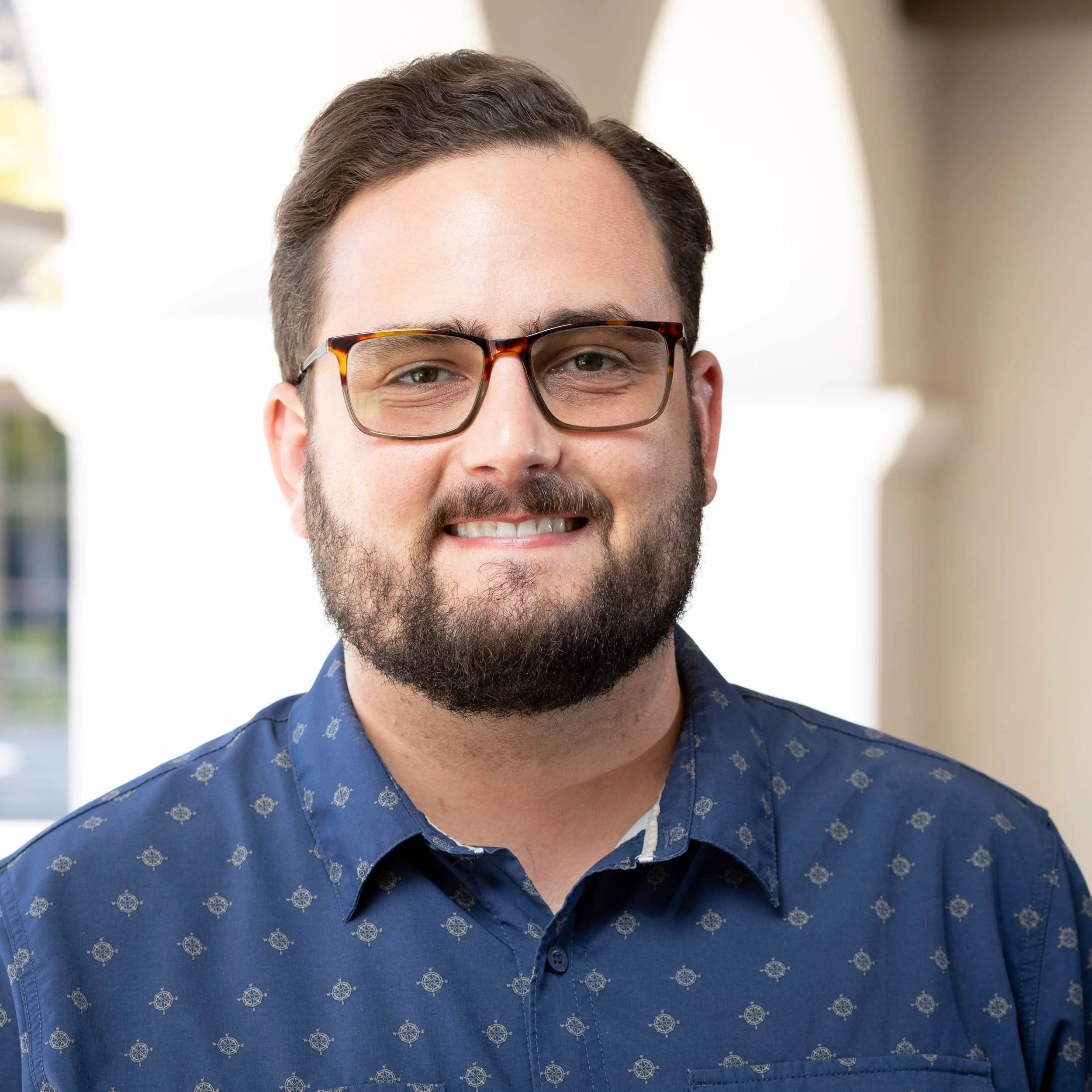 A man with glasses and a beard smiling, wearing a maroon polo shirt with a California Republic Leadership Academy logo, standing outdoors near a beige wall.