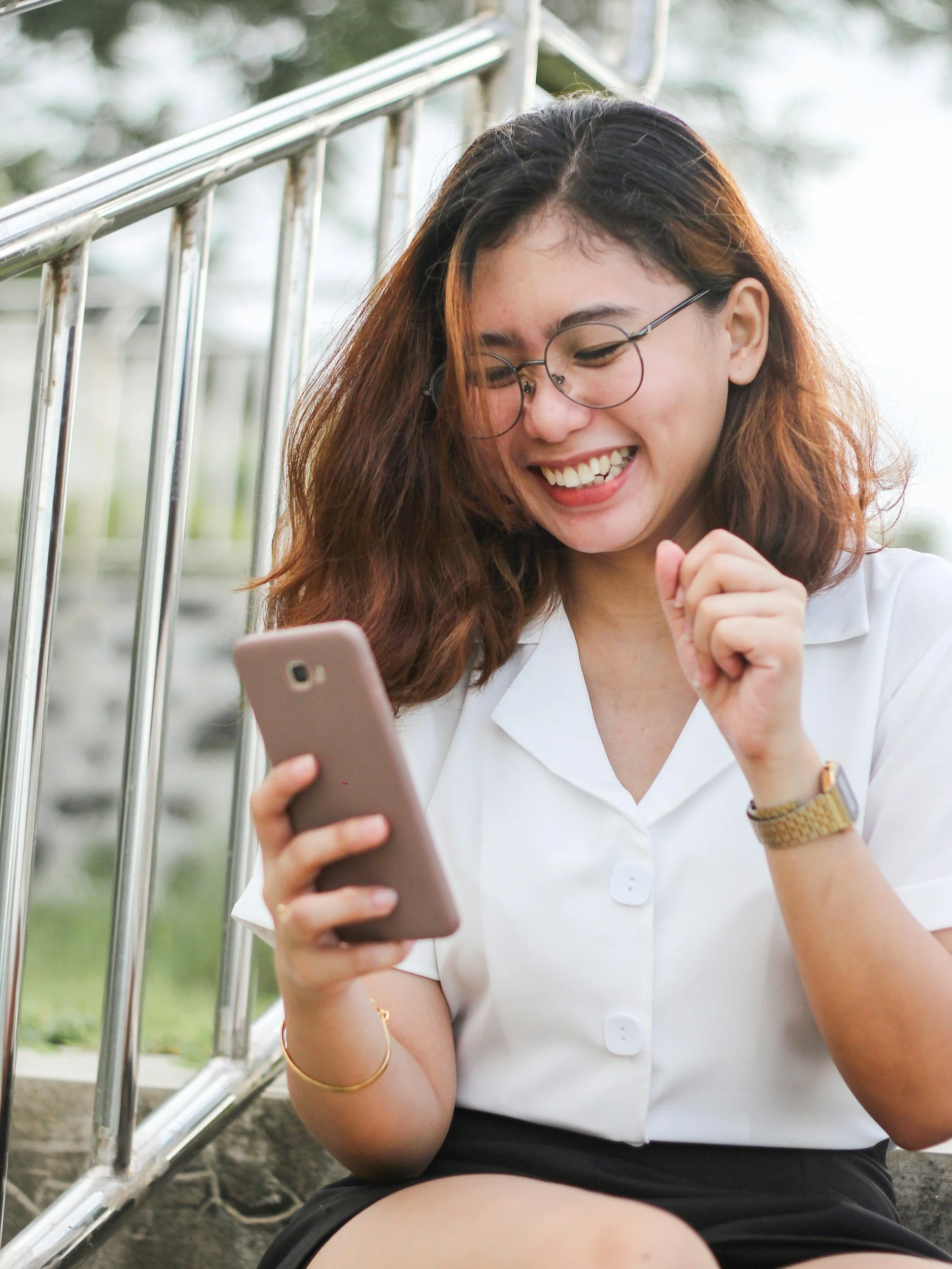 A young woman with glasses and wavy brown hair smiling as she looks at her smartphone outdoors