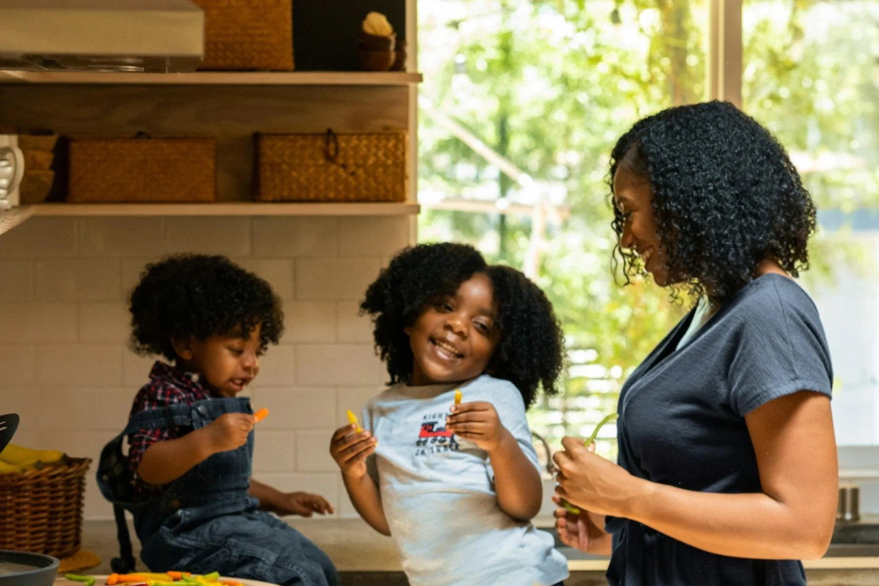 A woman and two young girls with curly hair enjoying a fun moment in a bright kitchen. The woman is smiling at the girl in the middle, who is holding a yellow toy, while the girl on the left is looking at a small orange object. The scene is illuminated with natural light coming from a window in the background.