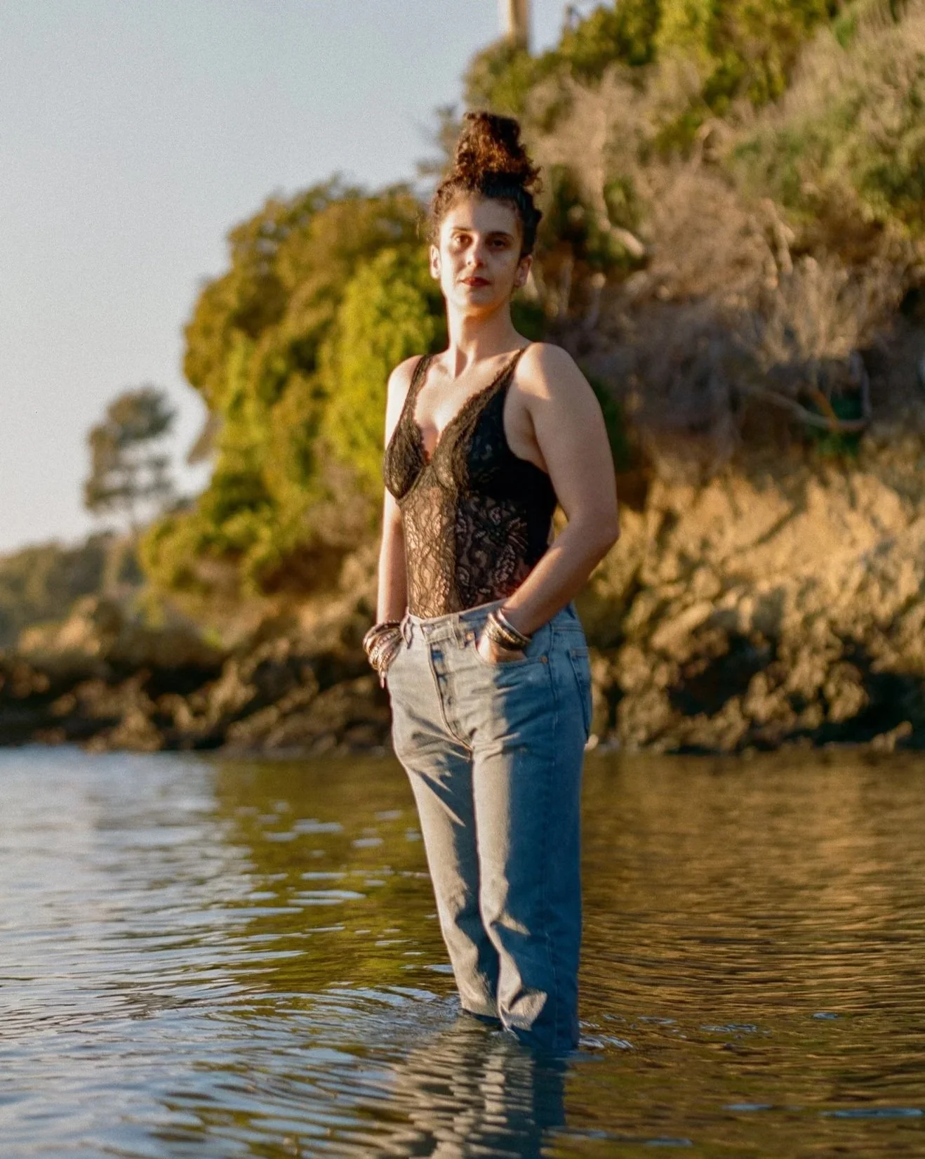 A full body photo of Shabnam Piryaei standing in water in a black lacy top and blue jeans