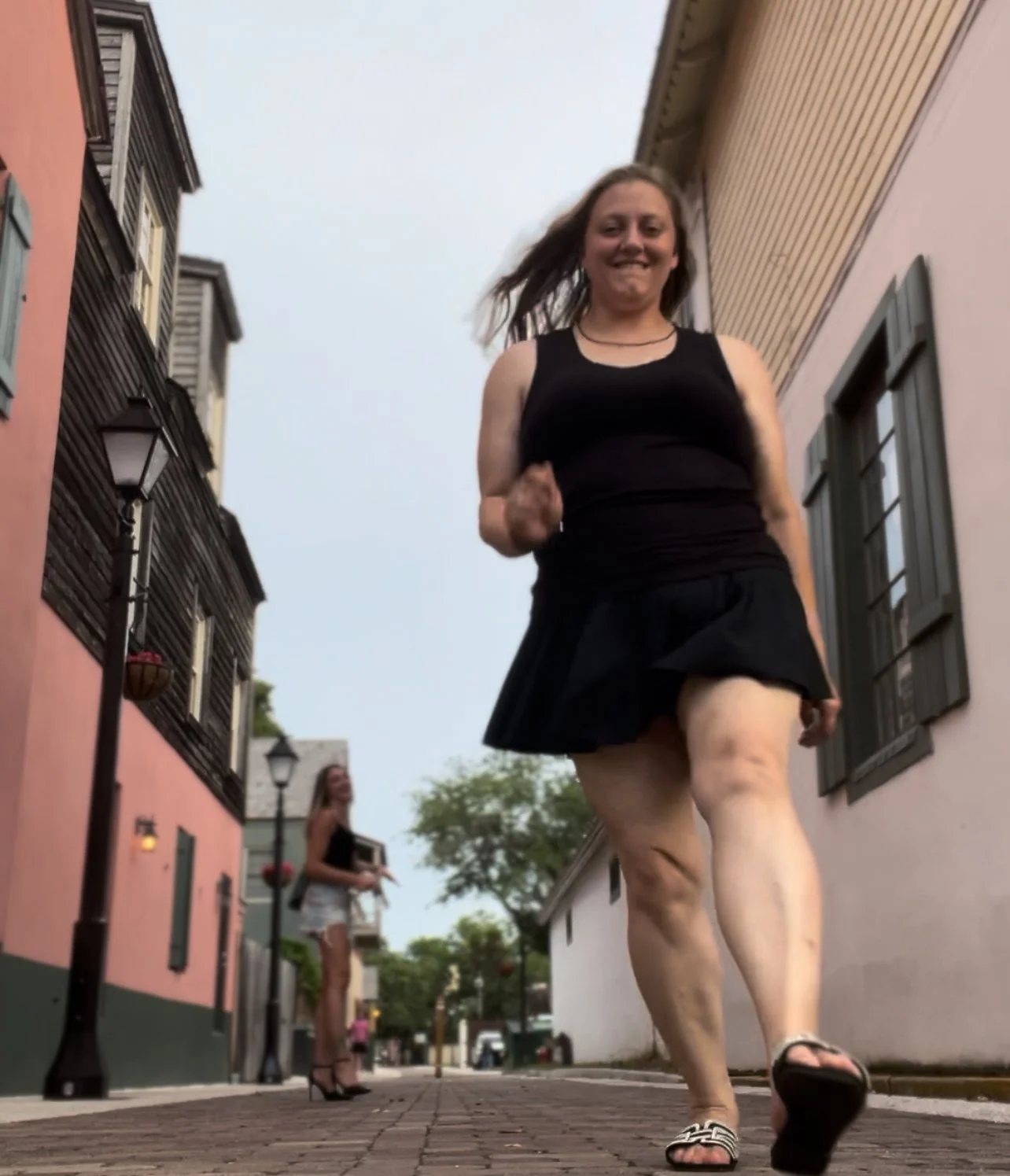 Amanda in a black tank top and black skirt skipping down the streets of St. Augustine, Florida. Pink and green buildings sit on to the left with aesthetic light poles placed perfectly along the street,