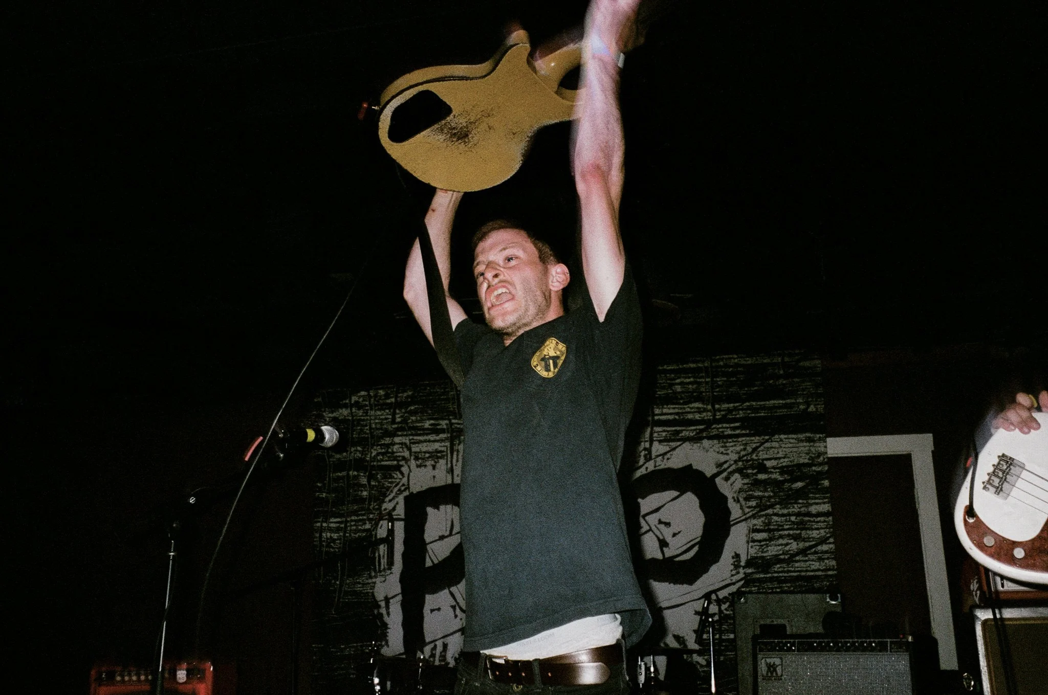 A man on stage raising a guitar above his head in an aggressive pose, with a black background and some musical equipment visible.