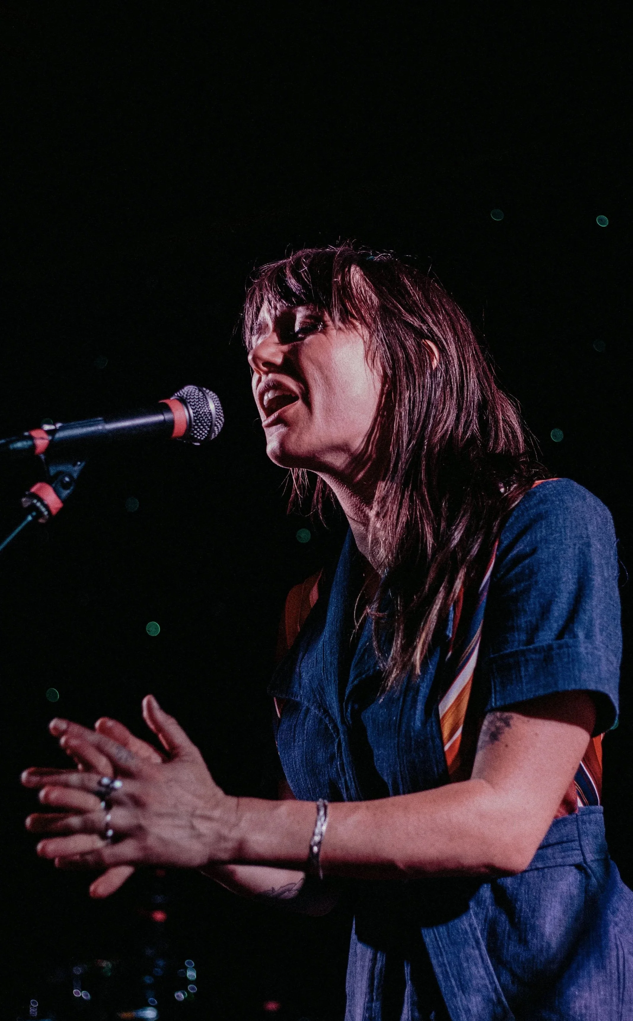 A woman singing passionately into a microphone on stage, wearing a denim shirt and shorts, with her hands clasped together.
