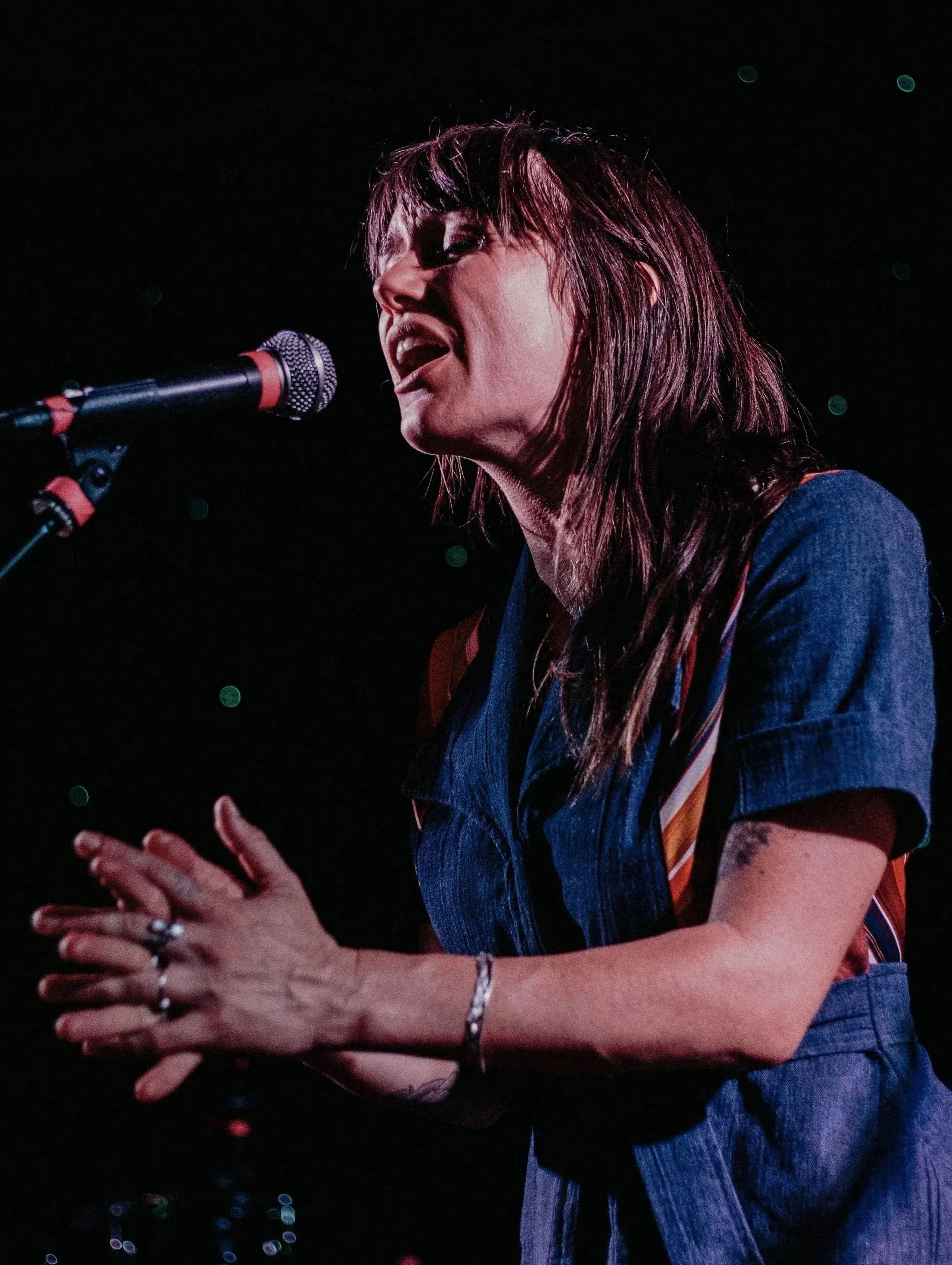 A woman singing passionately into a microphone on stage, wearing a denim shirt and shorts, with her hands clasped together.