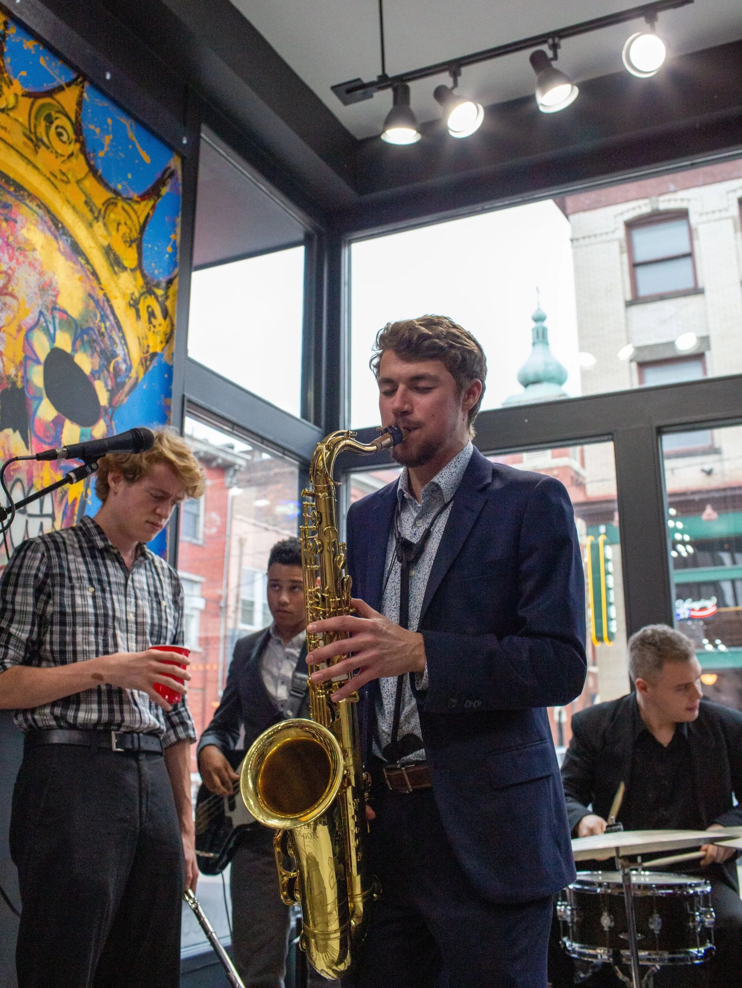 A young man in a suit playing a gold saxophone at an indoor event with other young musicians, some holding drumsticks and a drink, near large windows showing a city street and buildings.