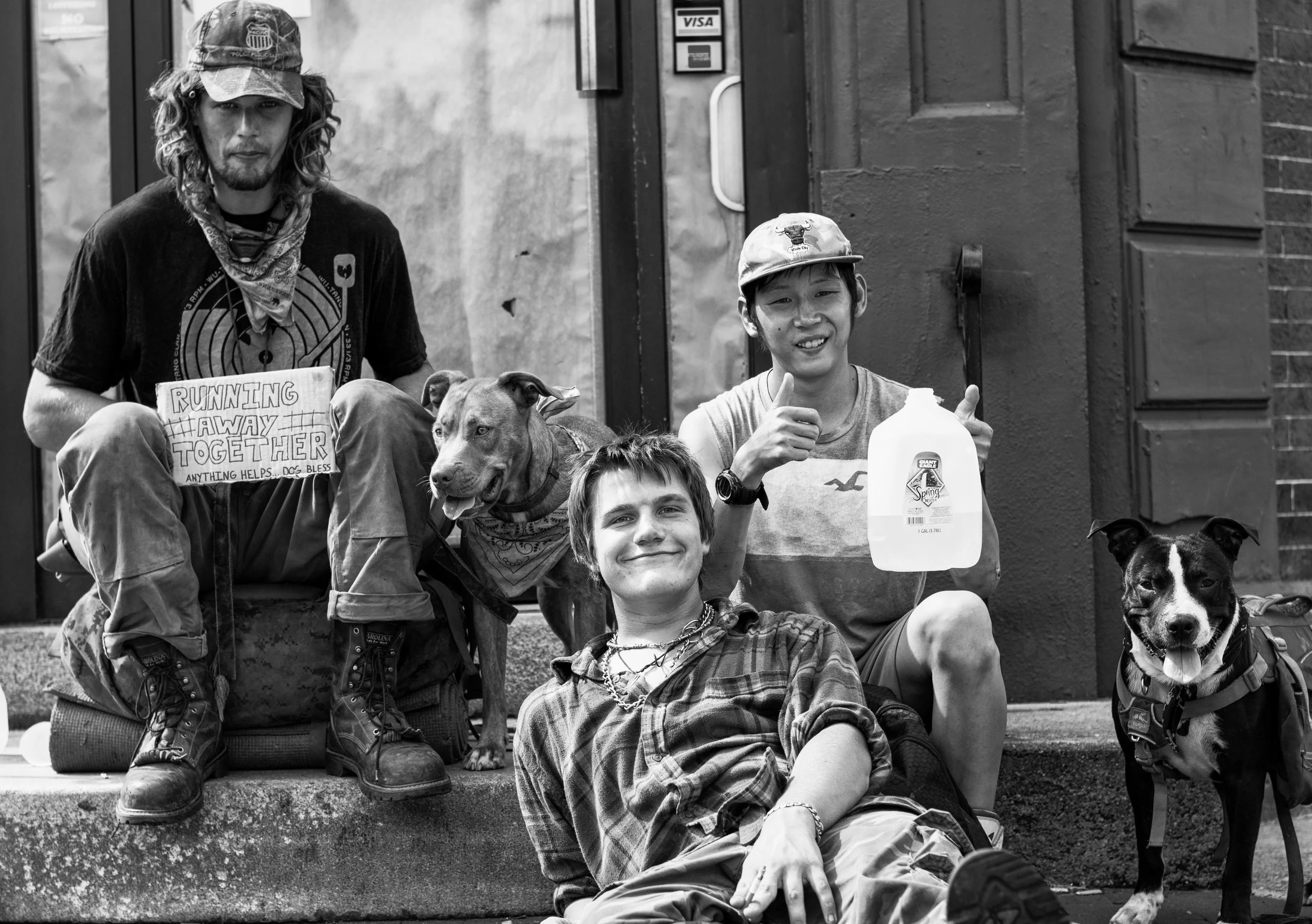 A group of three young men with two dogs sitting on a sidewalk outside a building. One man holds a sign reading 'Running the Way Together.' The man in the middle is smiling and giving a thumbs-up, holding a gallon of water. The third man is lying on 