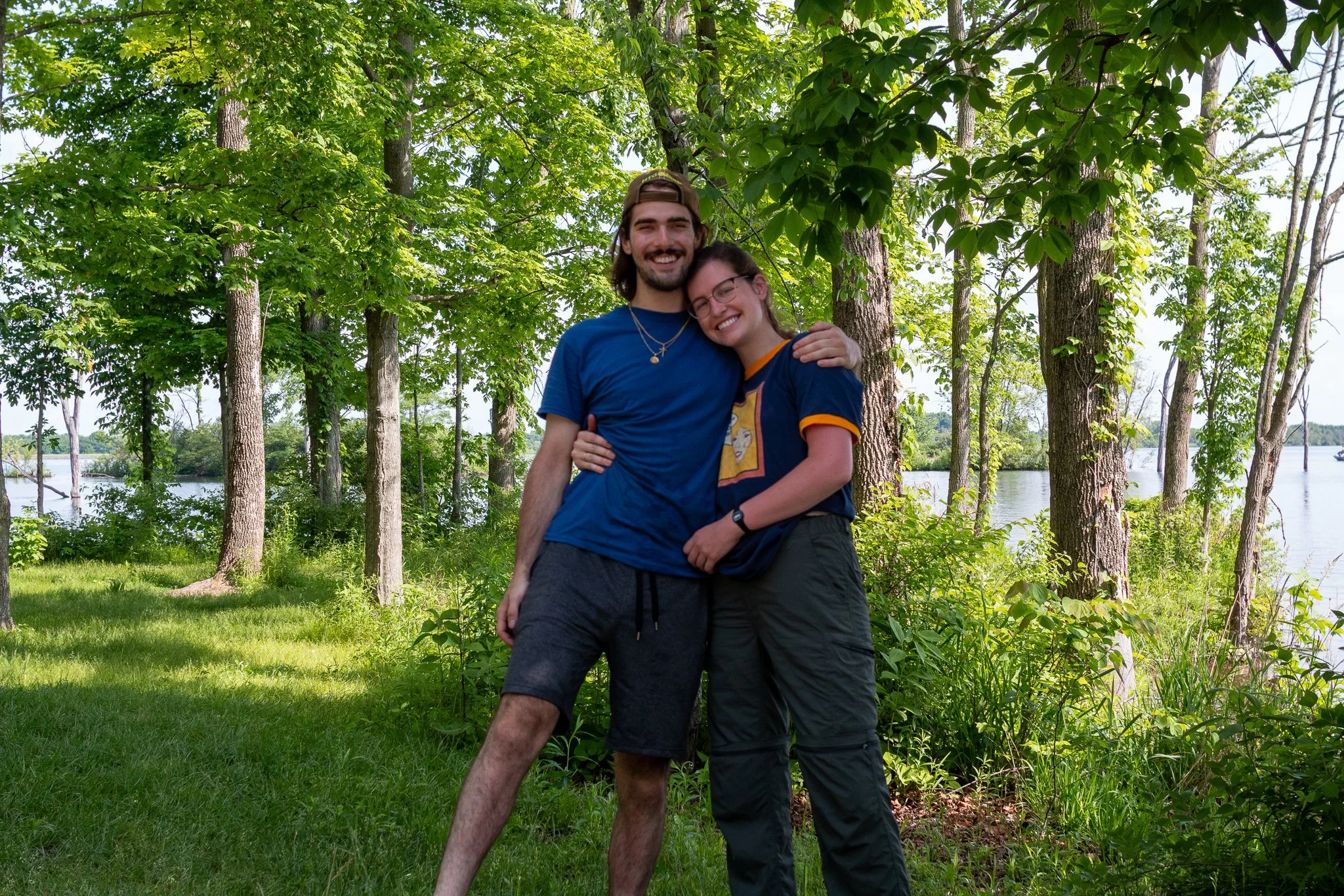 Two friends, a man and a woman, smiling and hugging each other outdoors in a green wooded area near water, with tall trees and bright sky in the background.