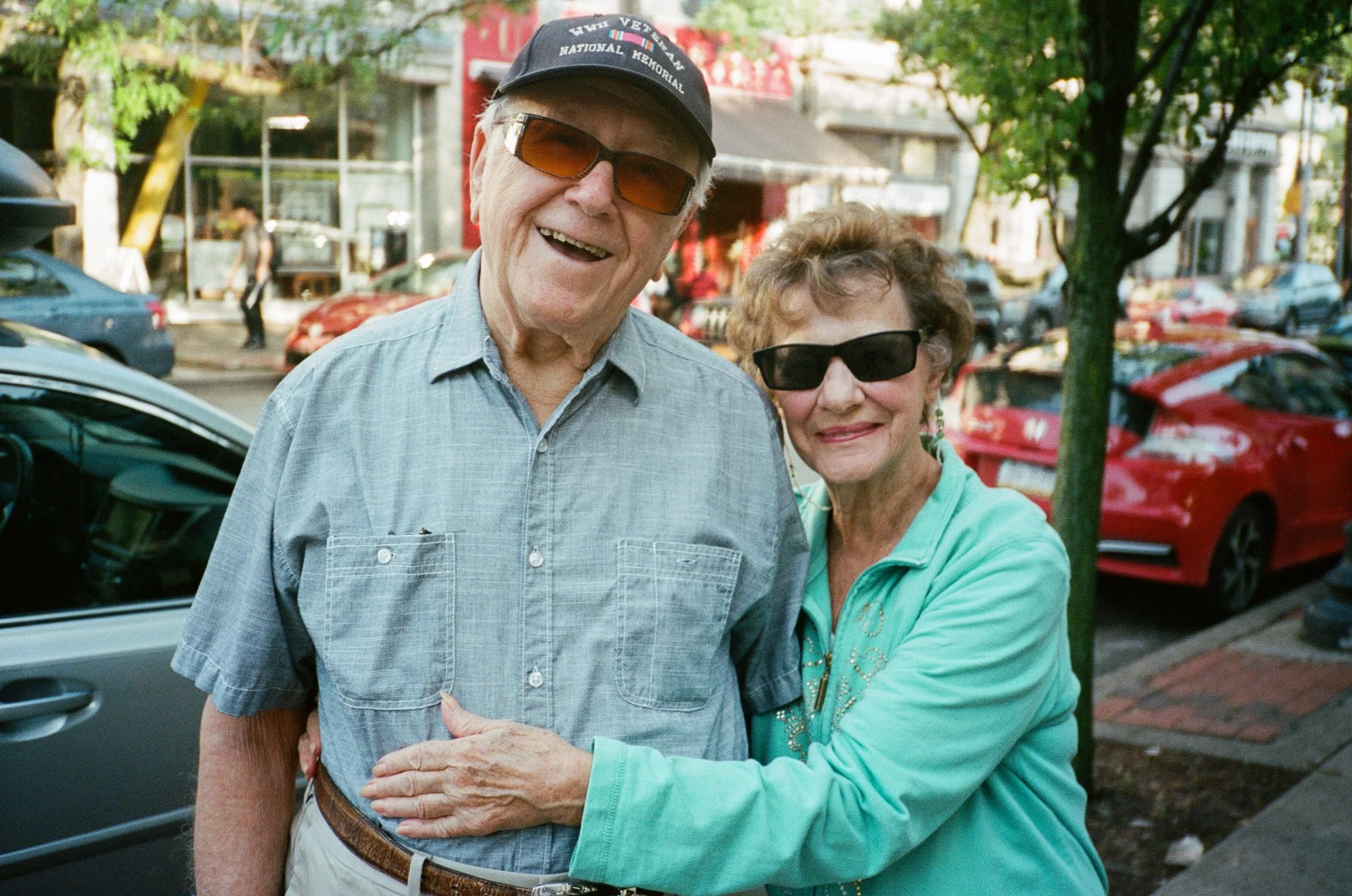An elderly man and woman smiling and wearing sunglasses outdoors on a city street, with parked cars and trees in the background.