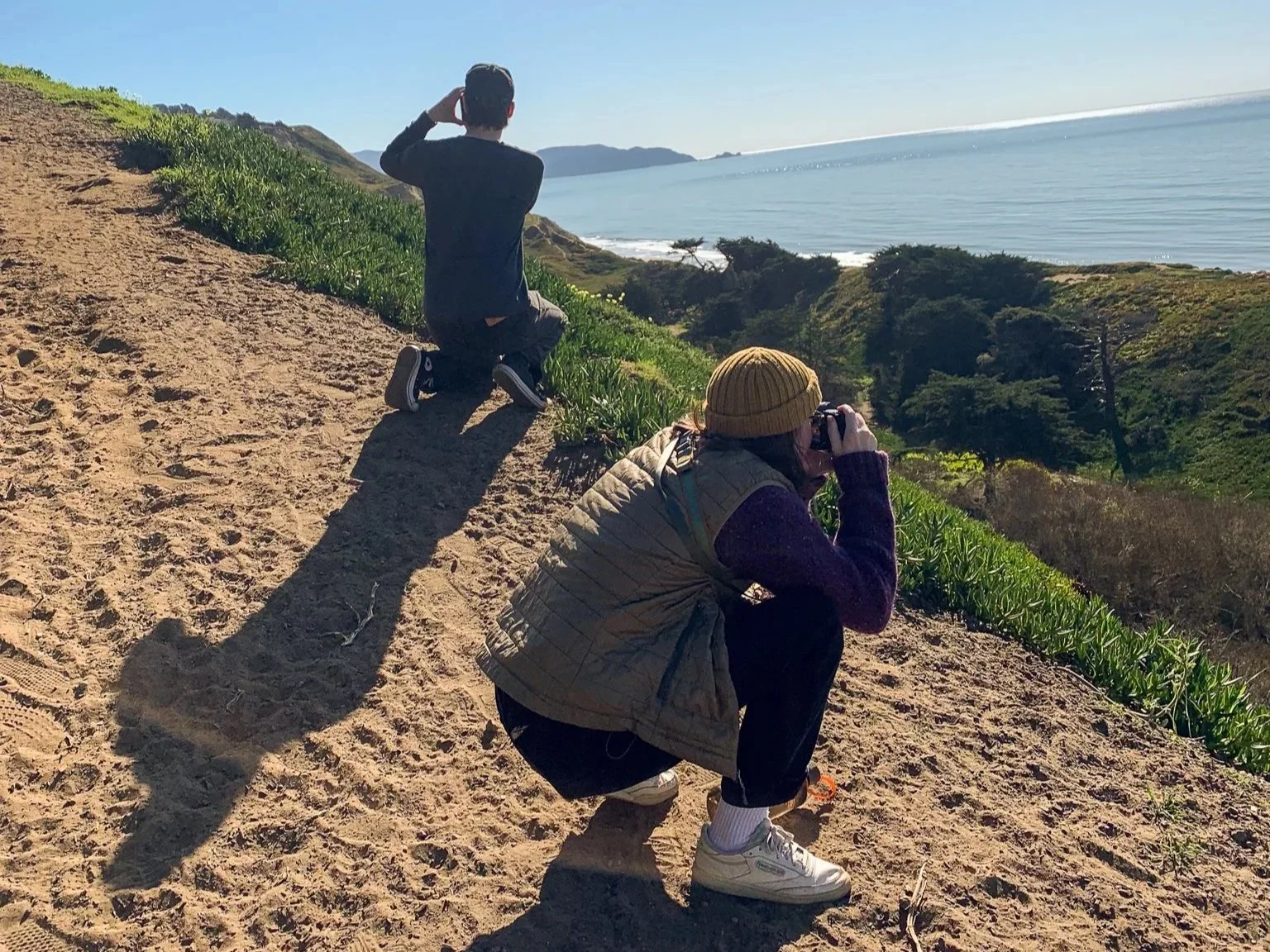 Two photographers on a sandy hillside near the coast. One is kneeling and taking pictures, the other is squatting and looking through binoculars or a camera. There are green plants and trees, with water and distant land in the background.