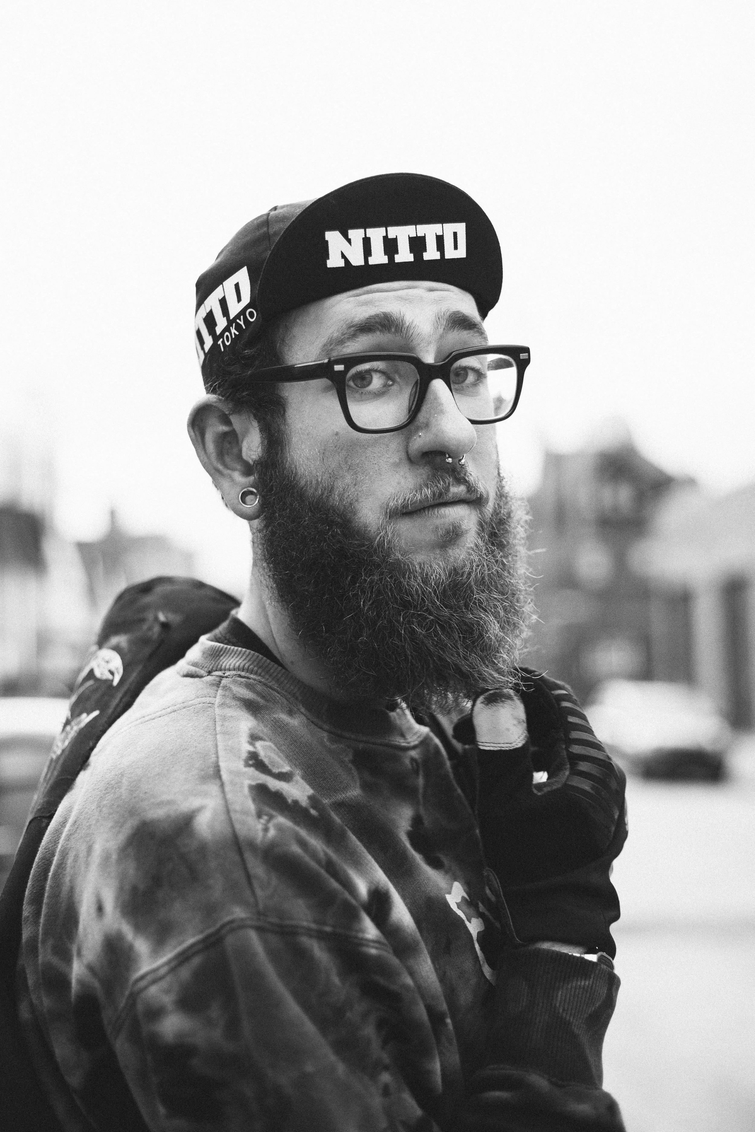 A young man with a beard and glasses wearing a cap with 'NITTO' written on it, a tie-dye shirt, and gloves, standing outdoors with buildings and cars in the background.