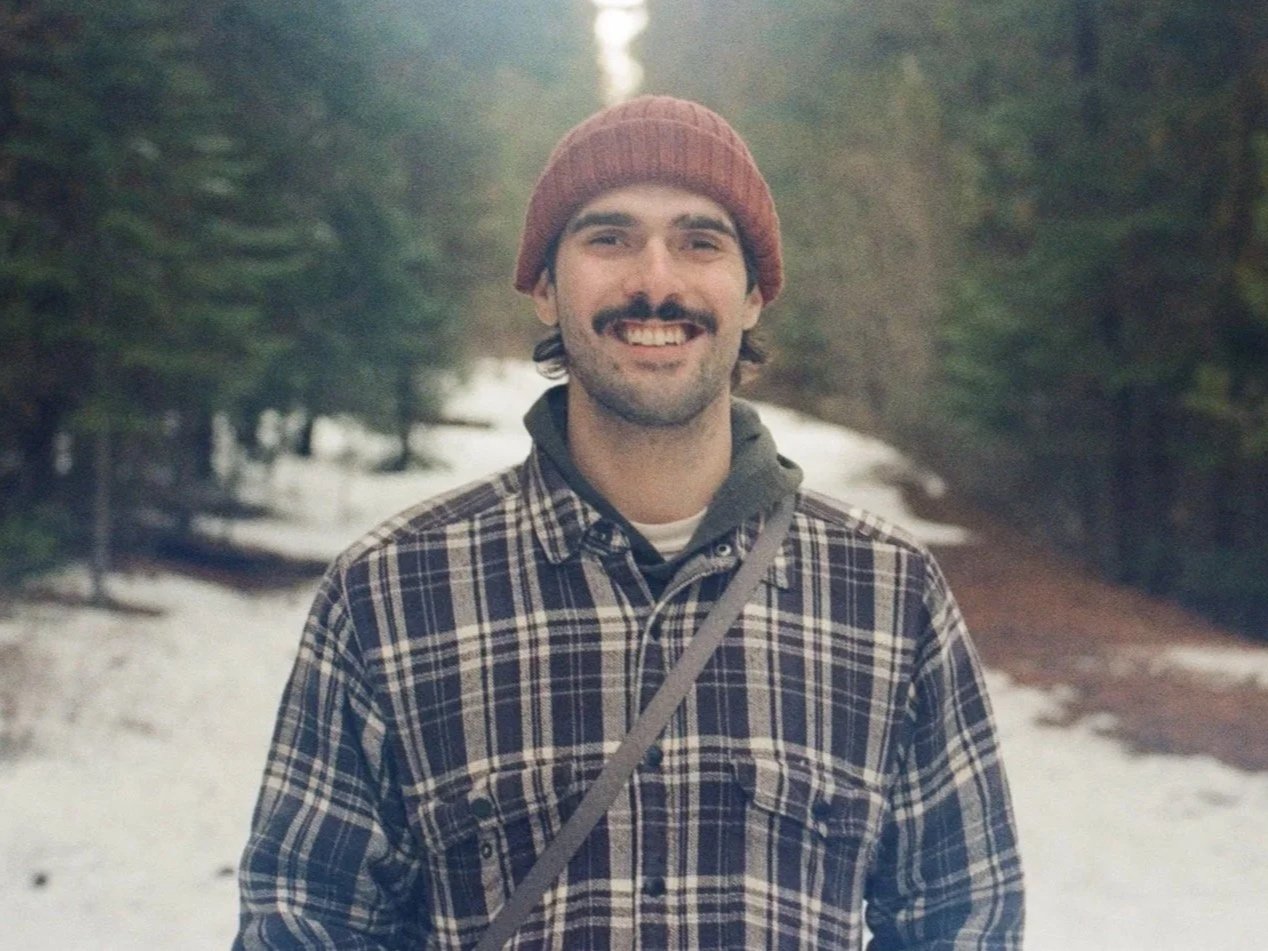 A man with a beard and mustache smiling outdoors in a snowy forest, wearing a red beanie, plaid shirt, and carrying a crossbody strap.