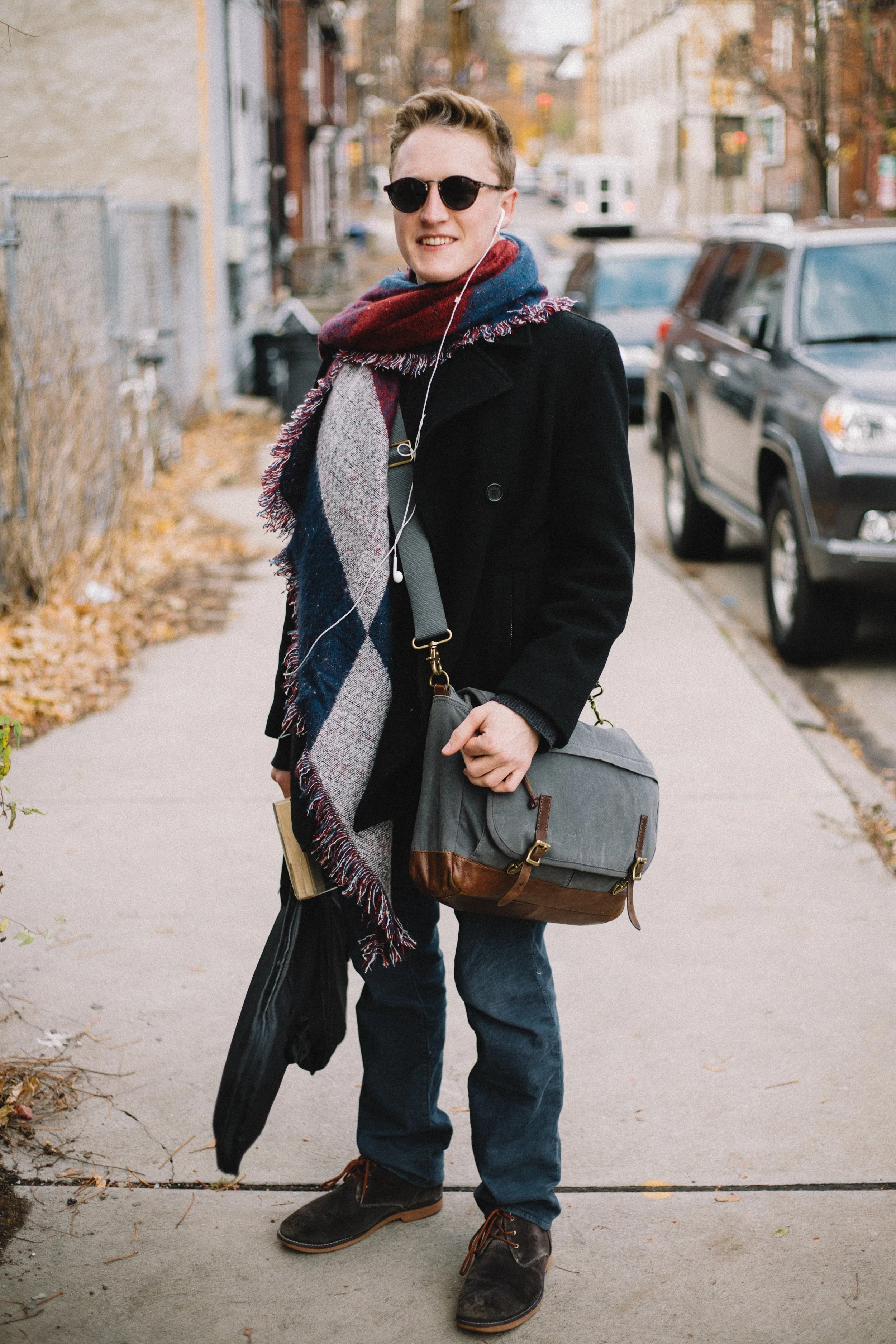 A young man standing on a city sidewalk, smiling, wearing sunglasses, a black coat, a multi-colored scarf, jeans, and brown shoes, holding a gray bag and an umbrella.