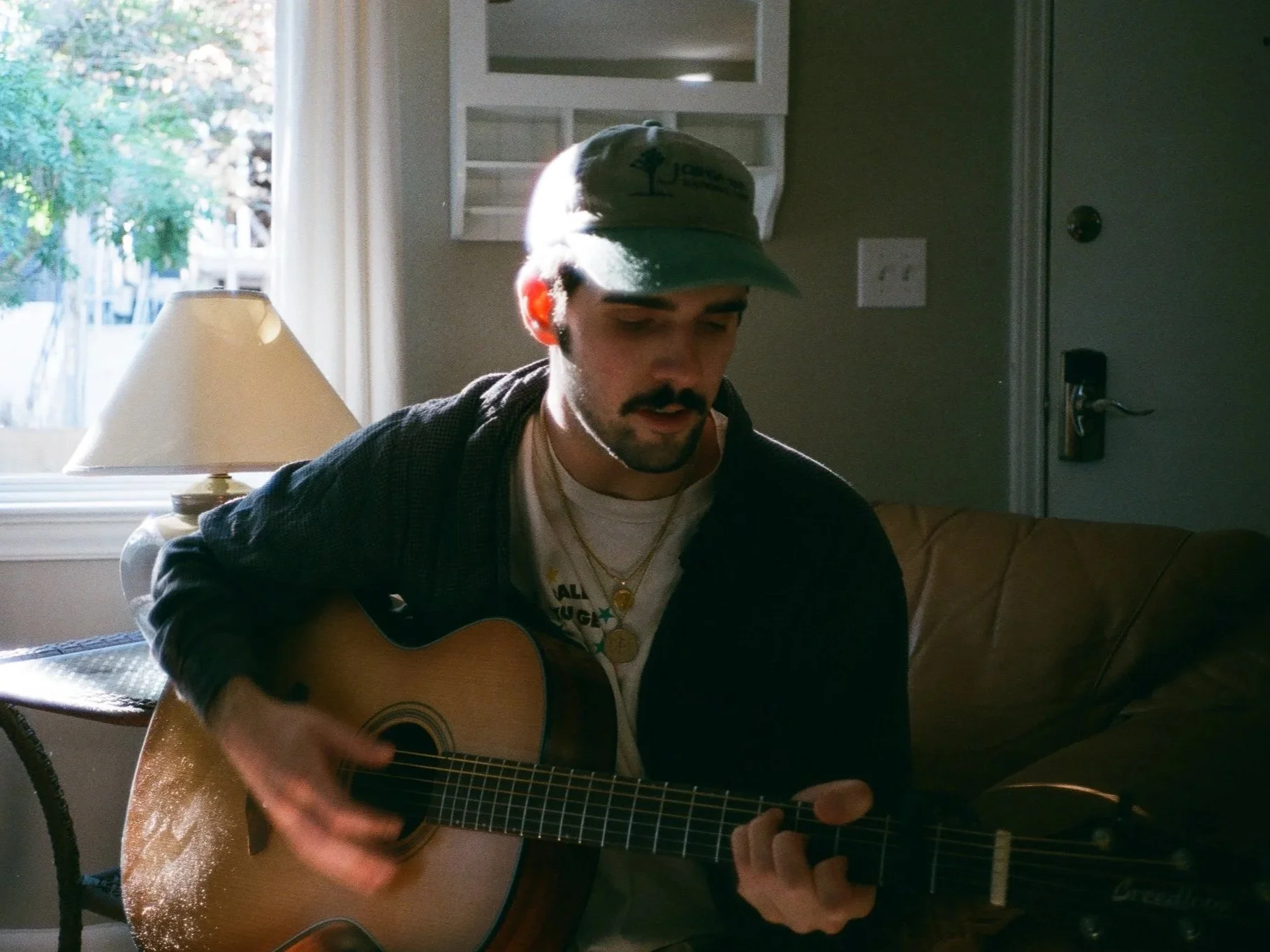 A young man with a mustache and dark hair, wearing a green cap and a layered necklace, is sitting on a couch playing an acoustic guitar. Behind him is a window with white curtains, and a table lamp is on his left. The background shows a beige wall with a small white box or shelf and a door to the right.