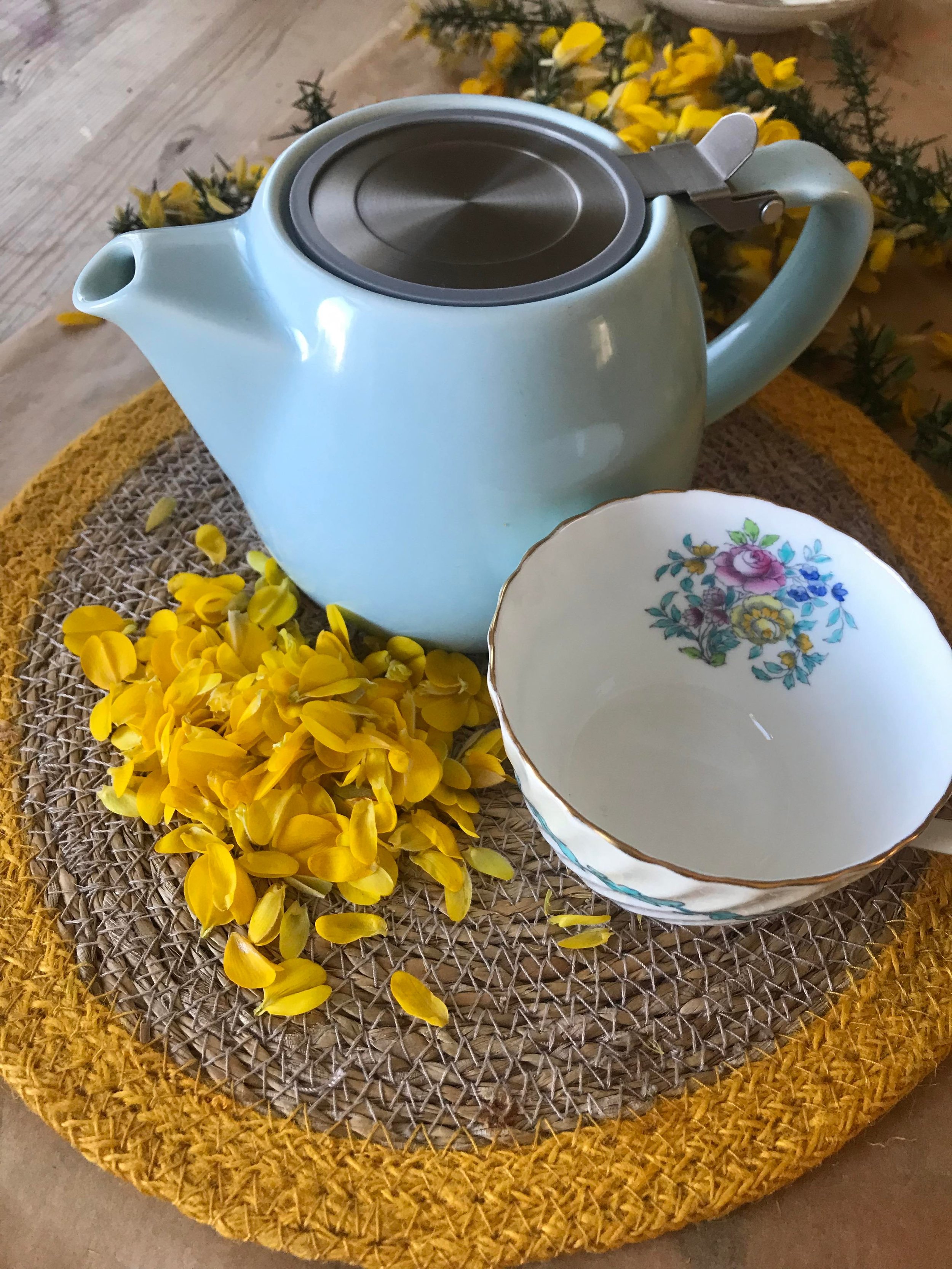 A light blue teapot with a patterned china cup sitting on a woven placemat edged with yellow, next to a pile of yellow gorse petals.
