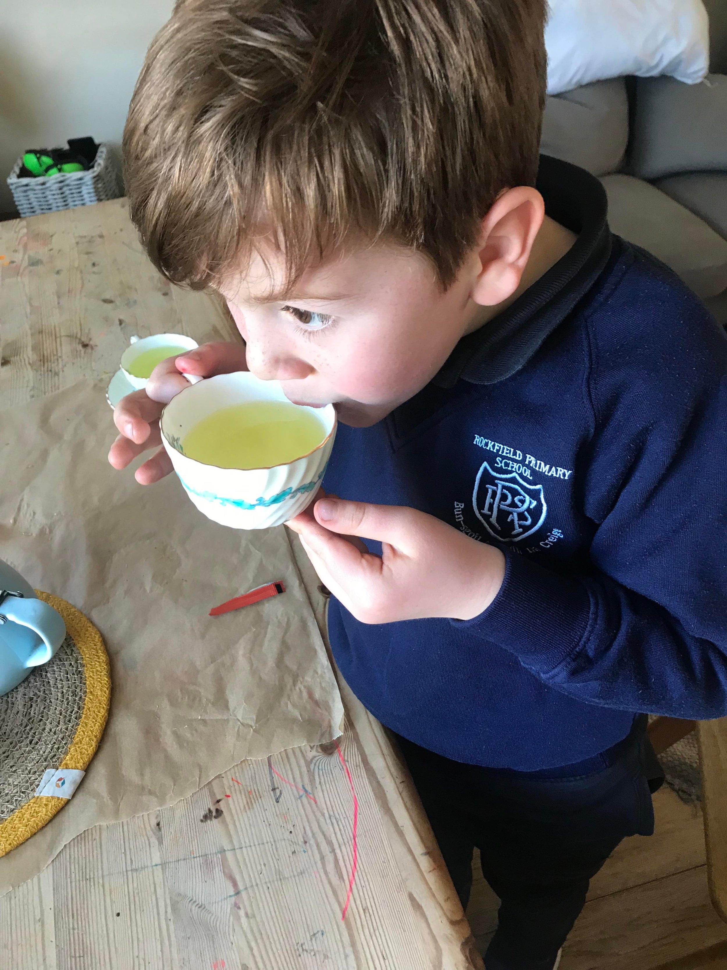 A boy of around seven sitting at a table sipping from a white teacup of pale yellow liquid. He is wearing a navy blue jumper which says Rockfield Primary School.