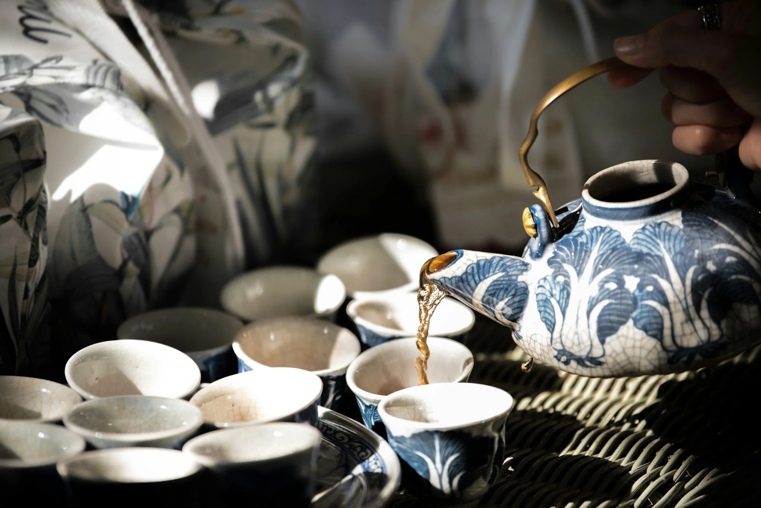 A hand pouring a dark liquid from a blue and white patterned teapot into small white cups arranged on a surface.