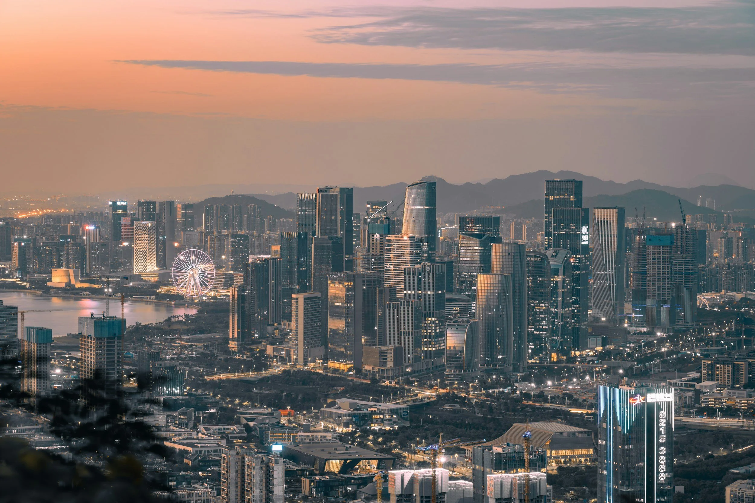 A city skyline during dusk with skyscrapers, a ferris wheel, and mountains in the background, illuminated by city lights.
