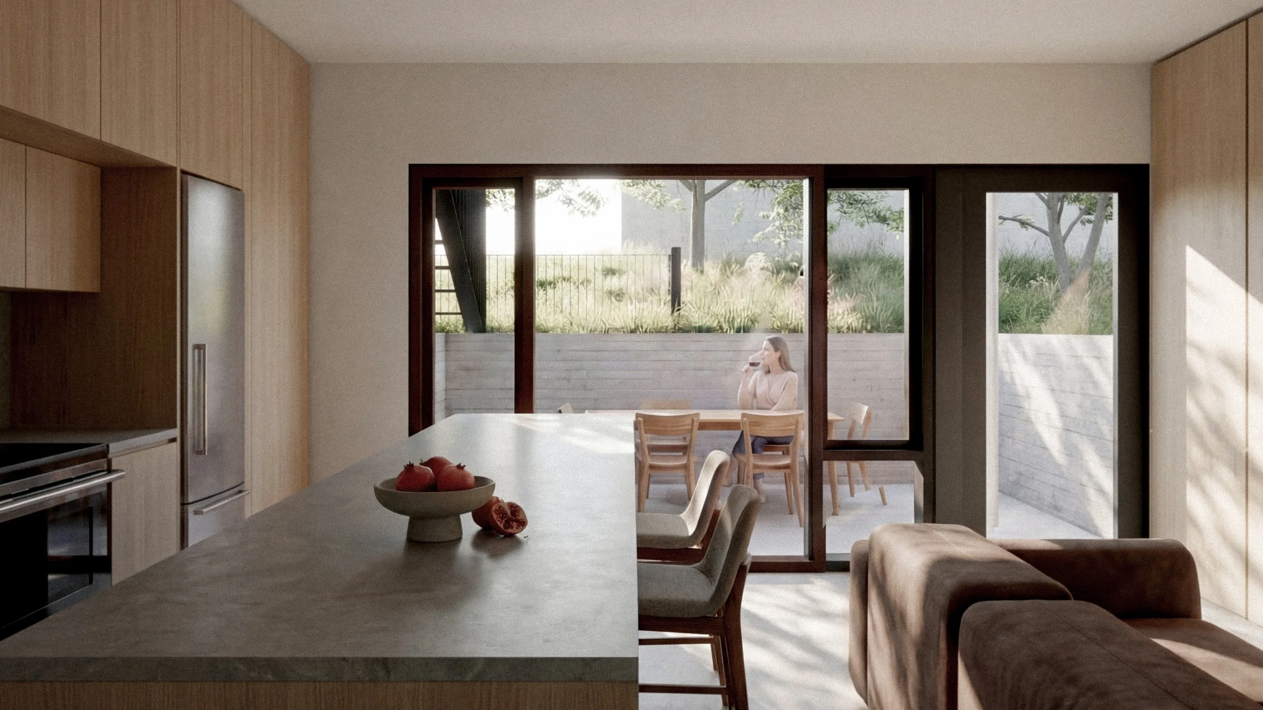 View of a modern kitchen with a large island, wooden cabinets, a refrigerator, and bar stools. Beyond a sliding glass door, a woman is sitting outside at a dining table, drinking a glass of wine, with trees and grass in the background.