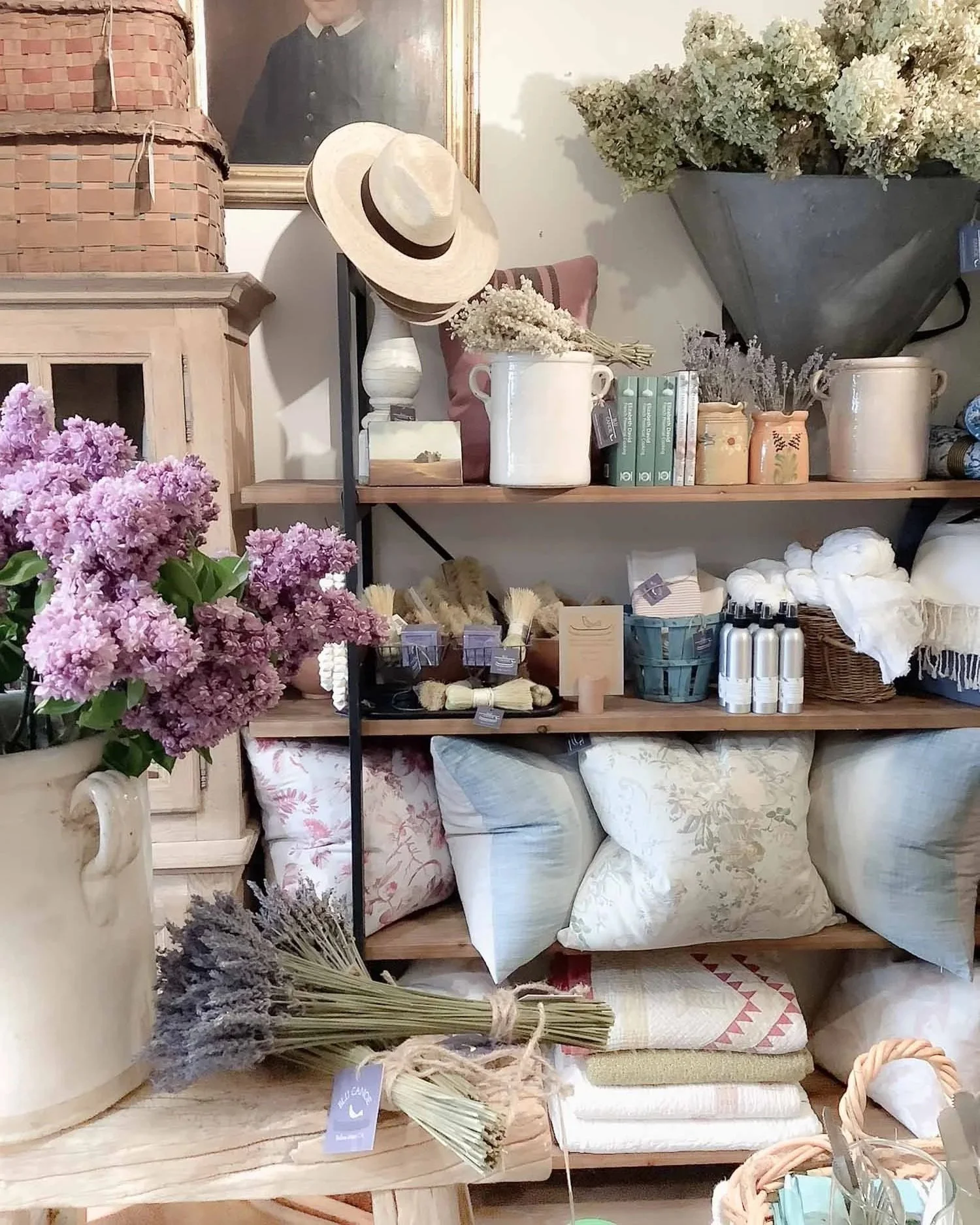 Display of decorative home items including a bouquet of purple hydrangeas, shelves with pillows, dried flowers, and various household decor in a store.