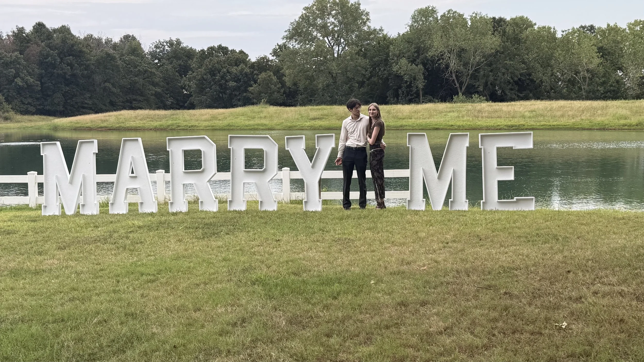 Ellie and Ricardo standing together in front of large “Marry Me” letters by the water during their engagement.