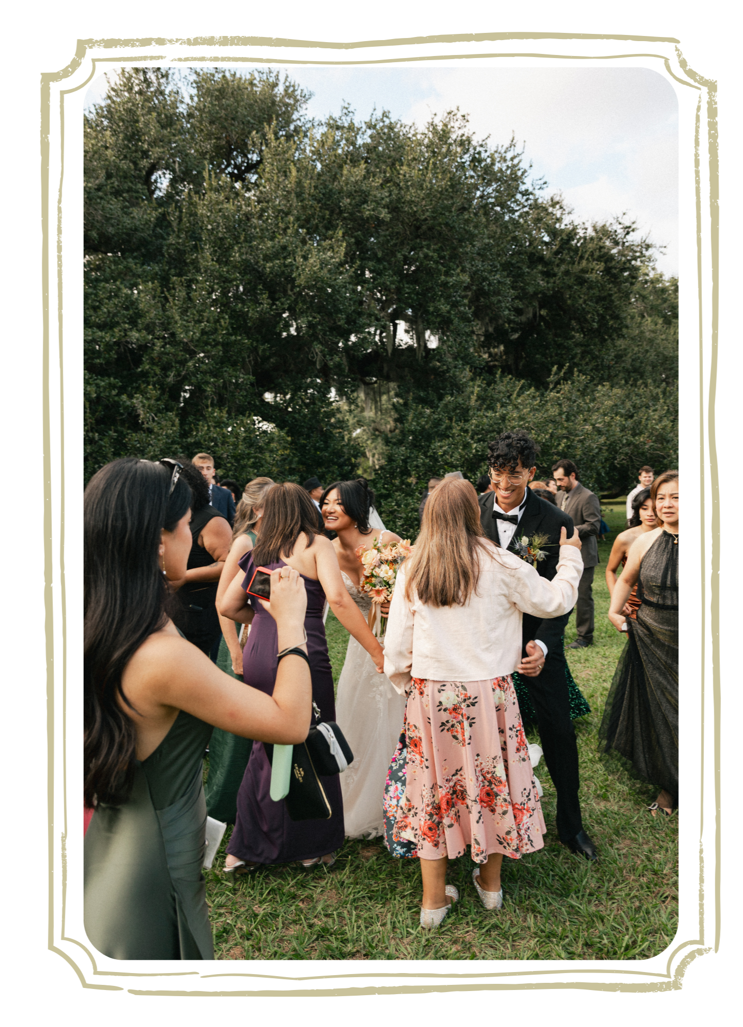 A group of people gathered outdoors on grass, dressed in formal attire, celebrating a wedding with trees and a cloudy sky in the background.