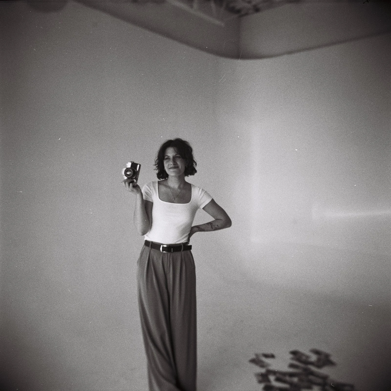 A woman with short curly hair, wearing a white top and striped pants, holds a vintage camera in her right hand, standing against a plain wall.