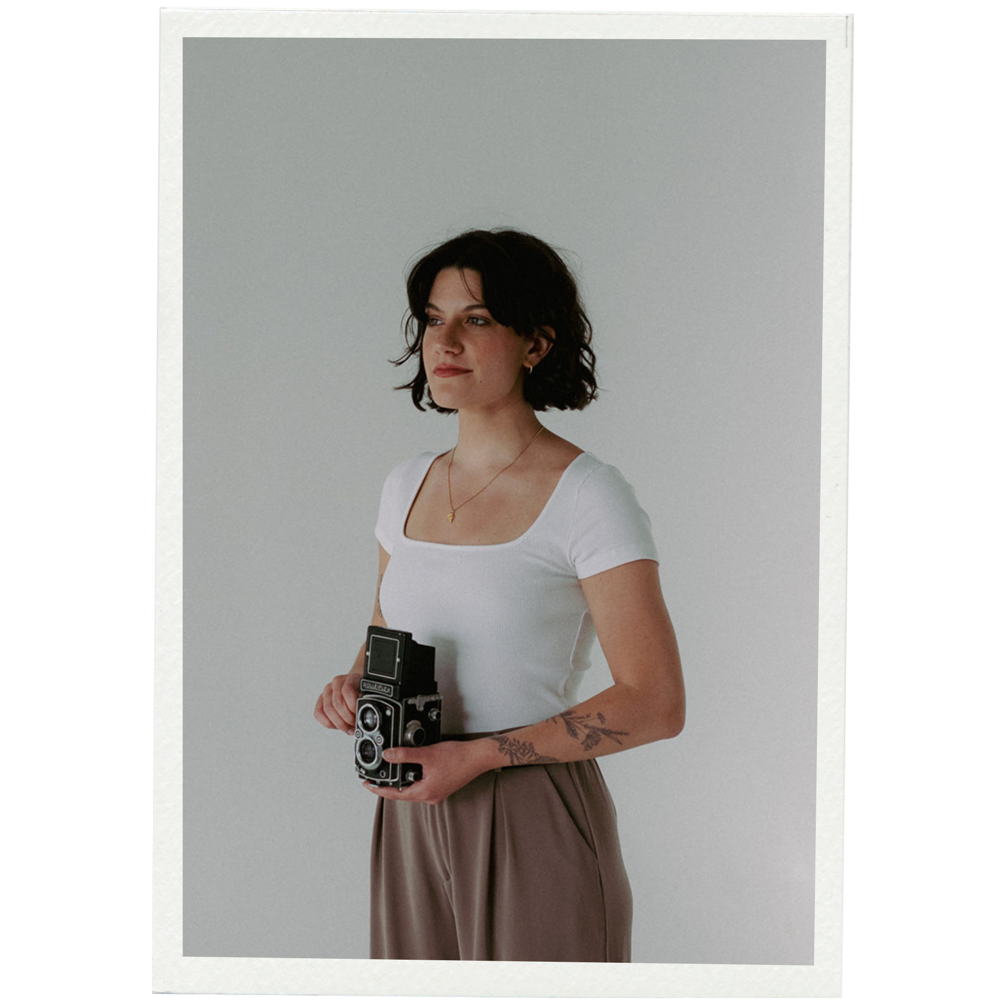A woman with dark hair and a necklace, wearing a white top and brown skirt or pants, holding a vintage camera, standing against a plain light gray background.