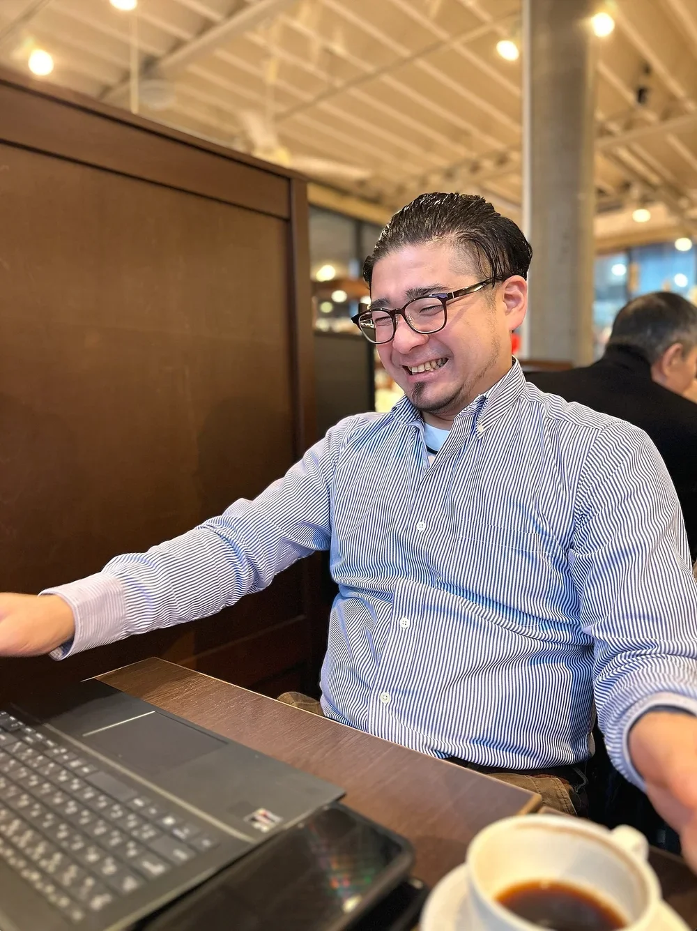 A man with glasses sitting at a table, smiling and laughing, with a laptop and a cup of coffee in front of him in a busy cafe or restaurant.