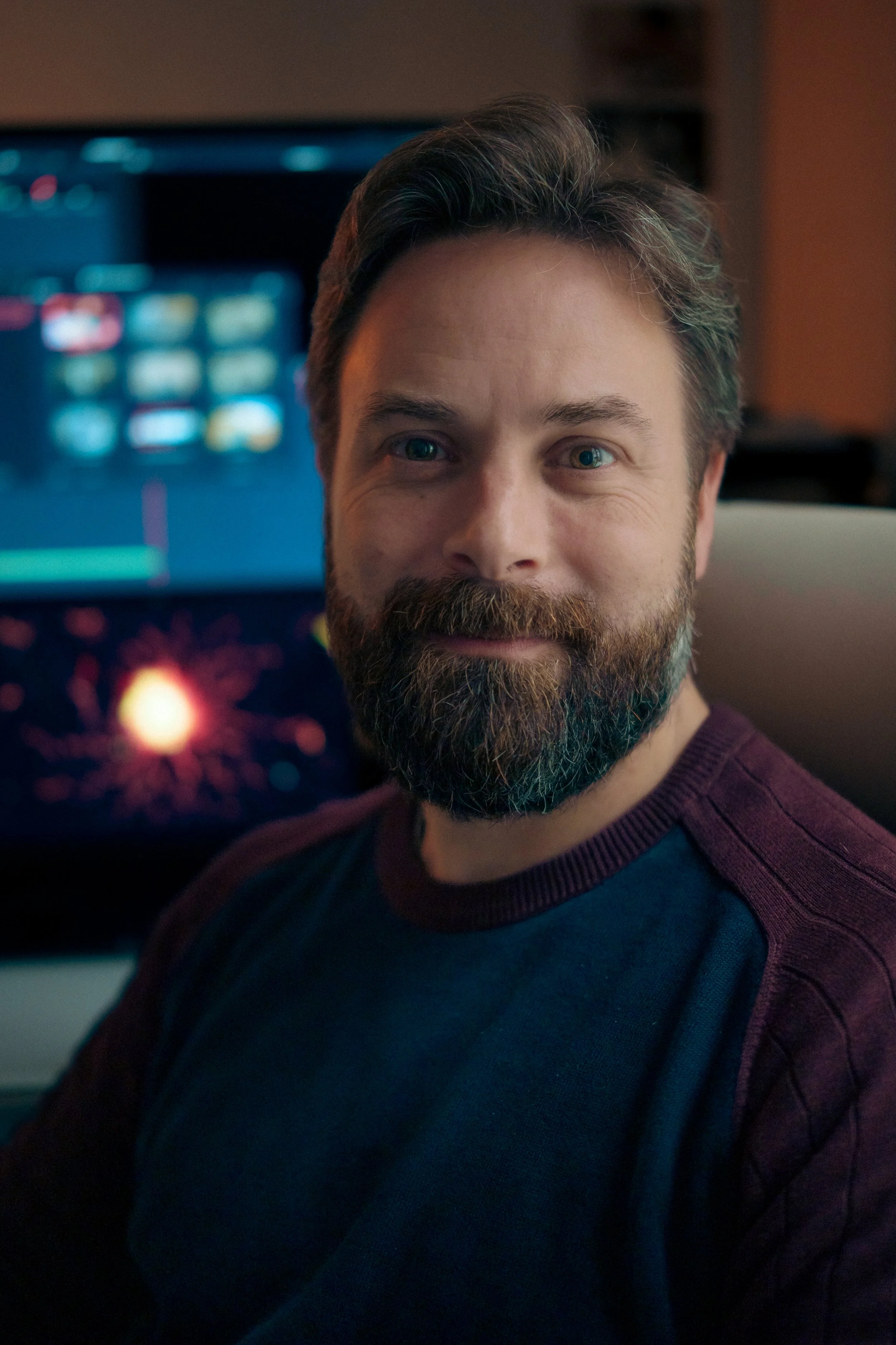 A man with brown hair and a full beard sitting in front of computer screens displaying colorful graphics and data, smiling at the camera.