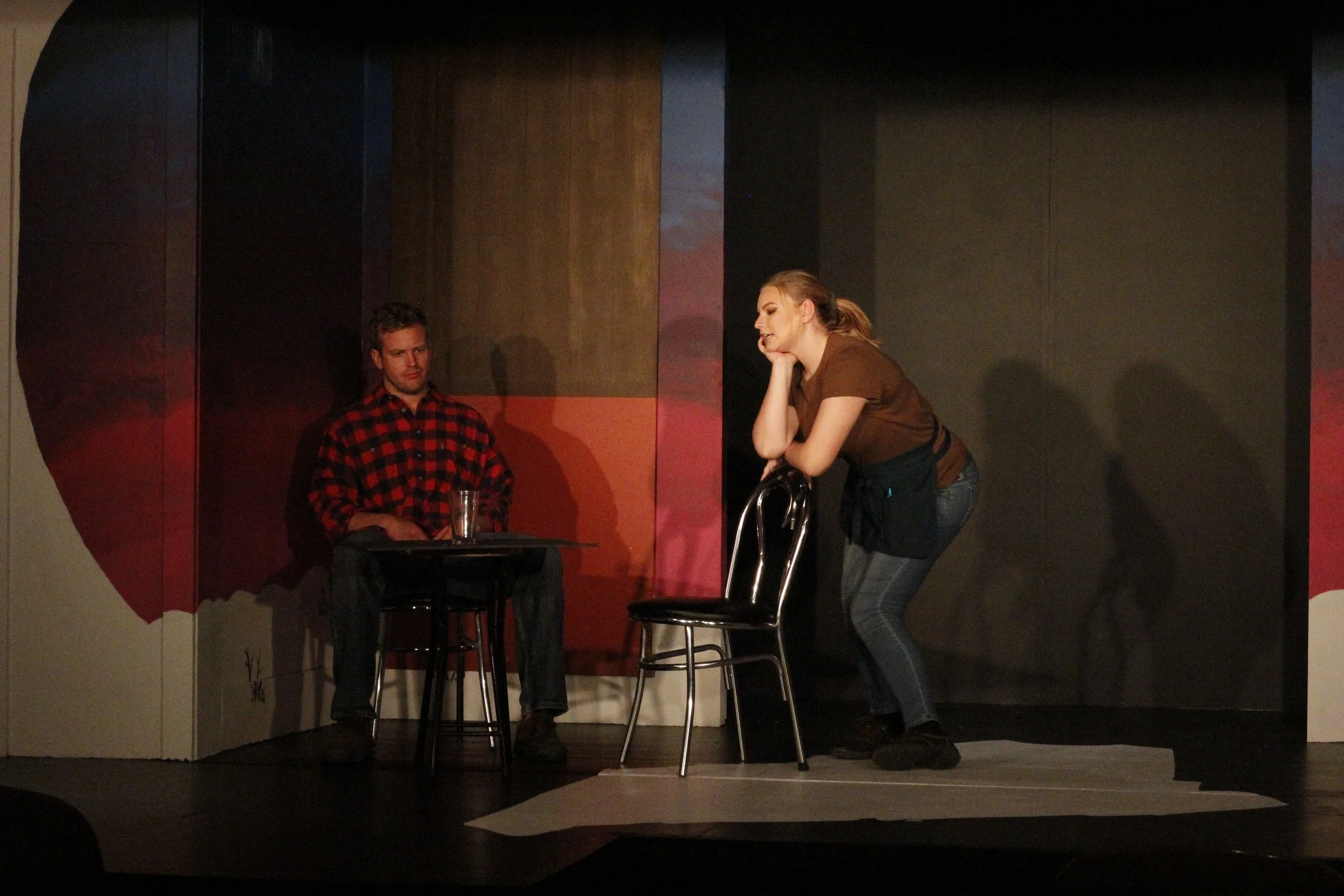 A man sitting at a table with a glass of water, and a woman leaning on a chair, appearing to perform in a play or comedy scene on stage.