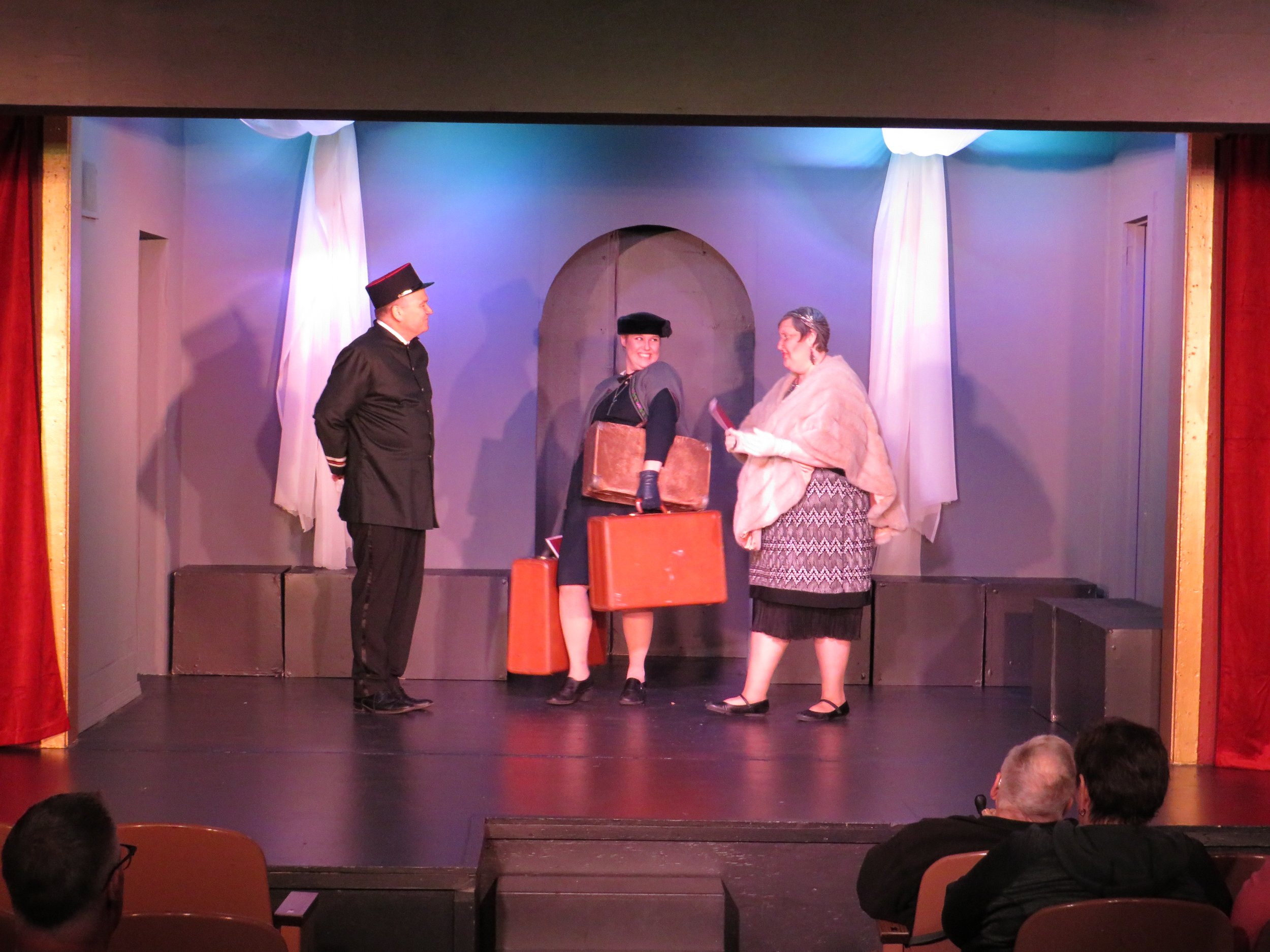A theatrical performance with three actors on stage, two women and one man, in period costumes, with an audience watching.