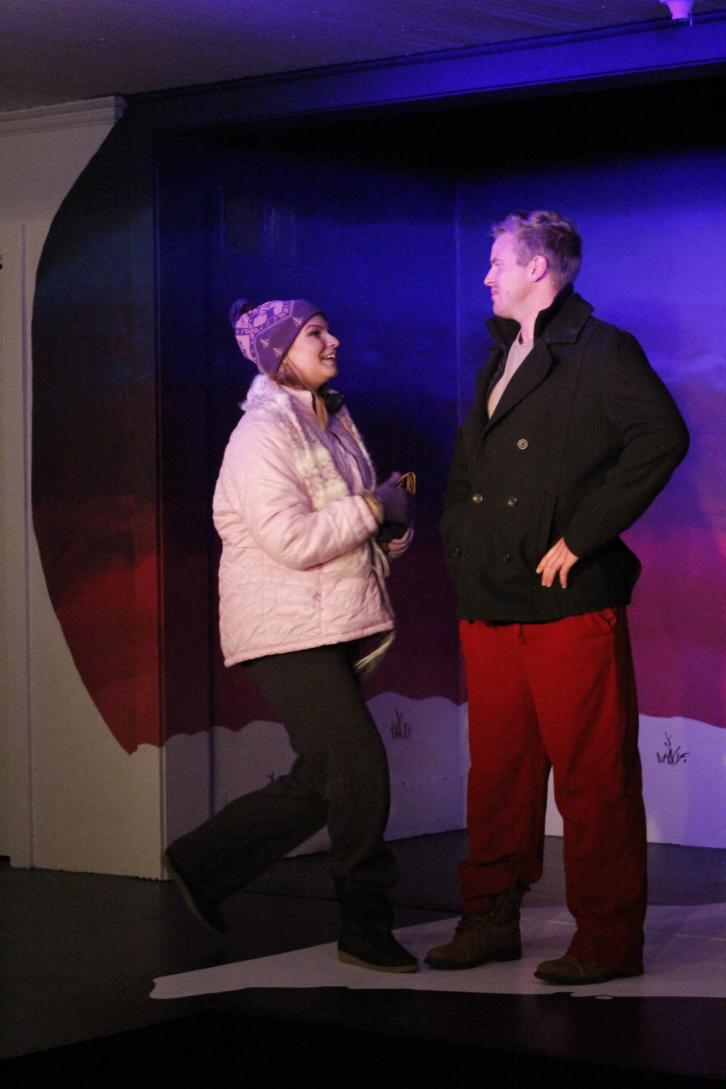 A woman and a man are having a conversation, standing on a paper cutout of snow with a painted winter scene behind them. The woman is wearing a pink quilted jacket, a knit hat, and gloves, and the man is wearing a black coat and red pants.