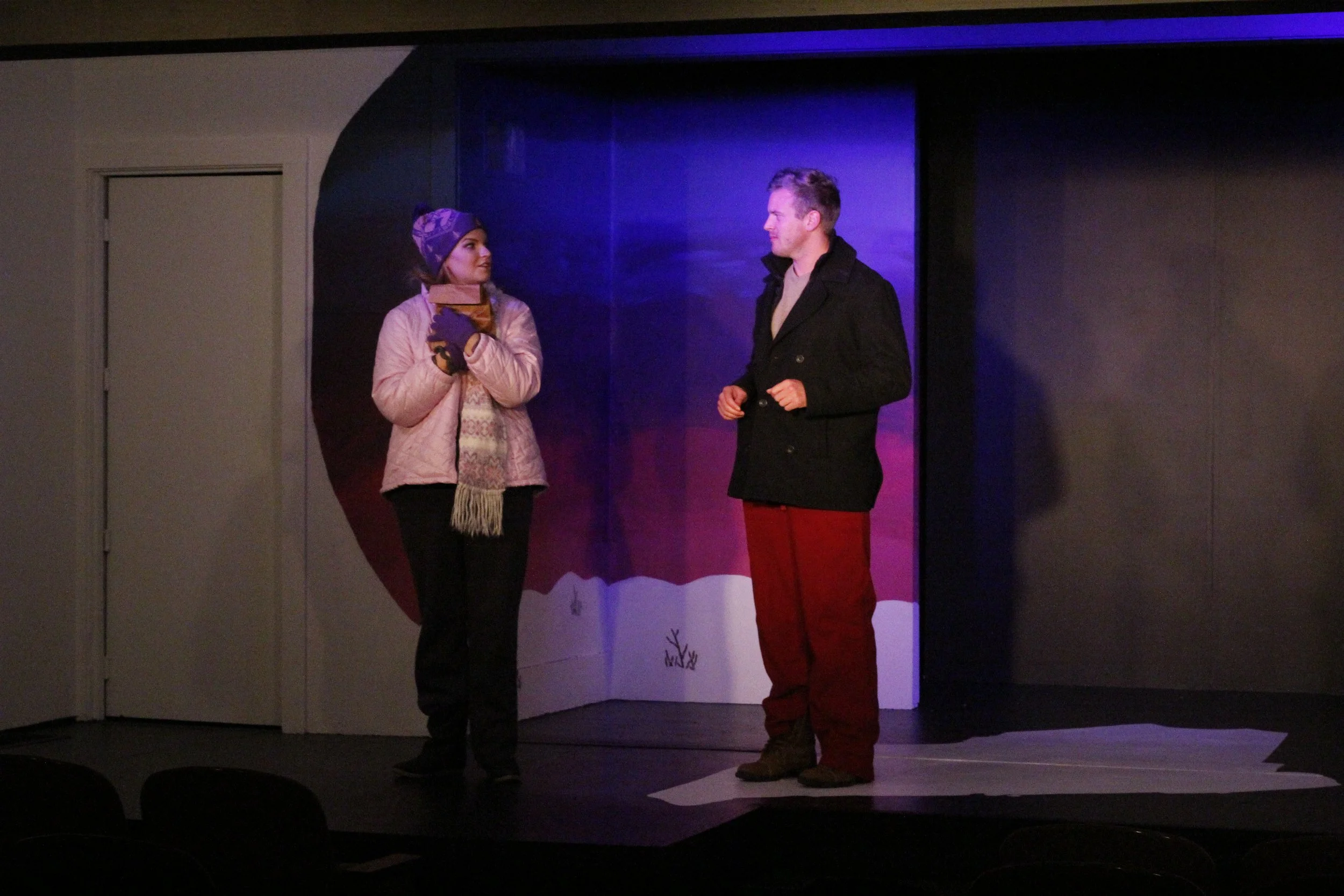 Two people, a woman dressed in winter clothing and a man in a black coat, stand on a stage with a colorful set behind them, engaged in conversation.