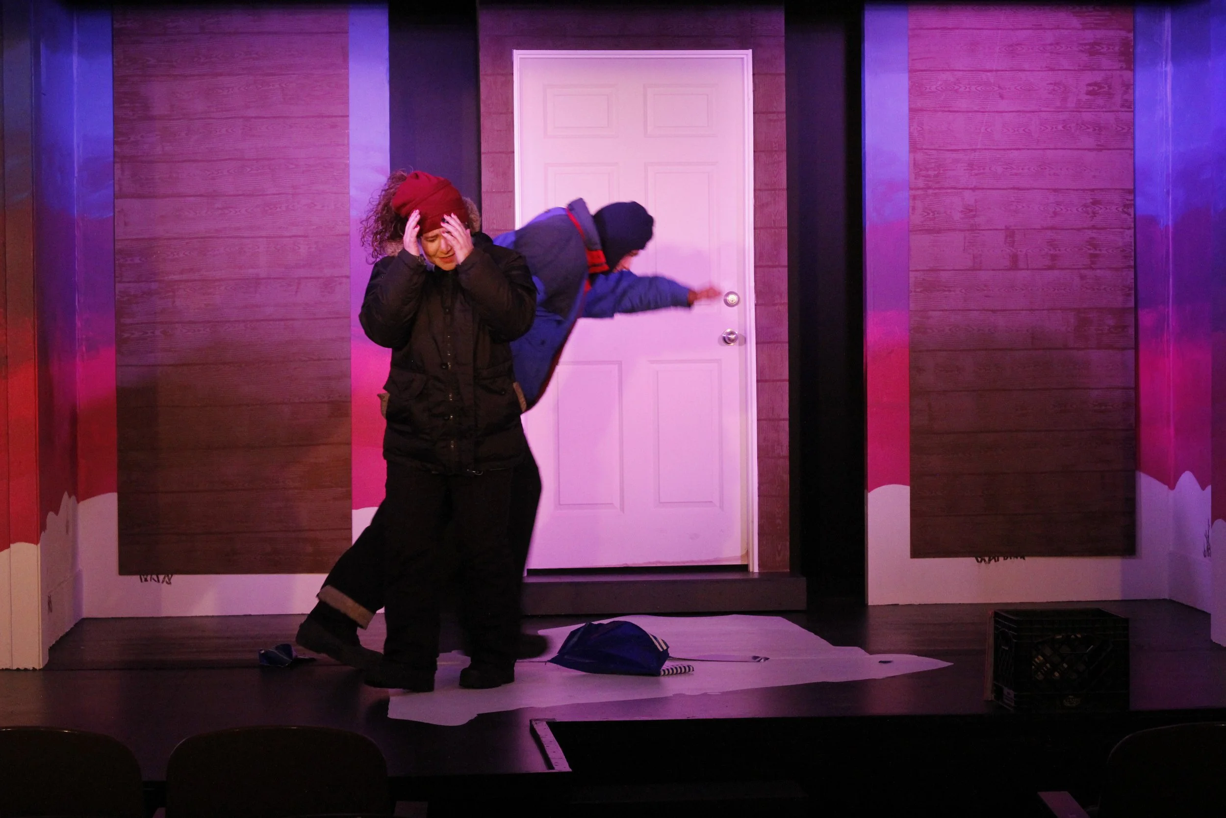 Two actors performing a scene on stage, one woman with curly hair and a red hat, holding her head, and one man bent over reaching towards a door, set against a pink and purple background.
