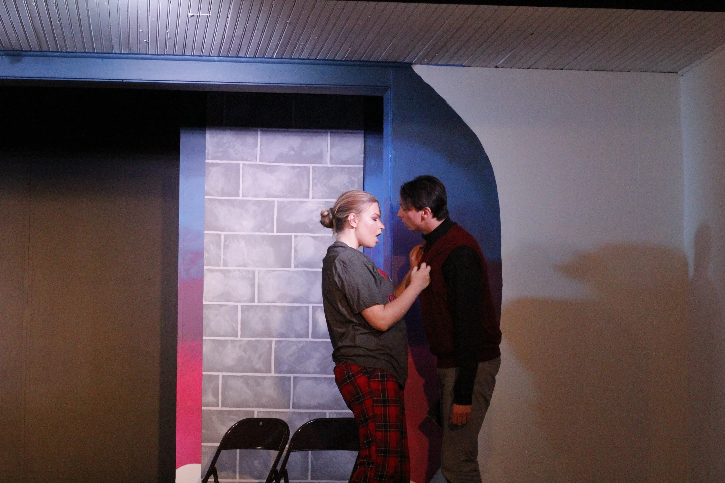 A man and a woman standing close together, with the woman touching the man's chest, in front of a blue and gray wall. There are two black chairs at the bottom of the scene.