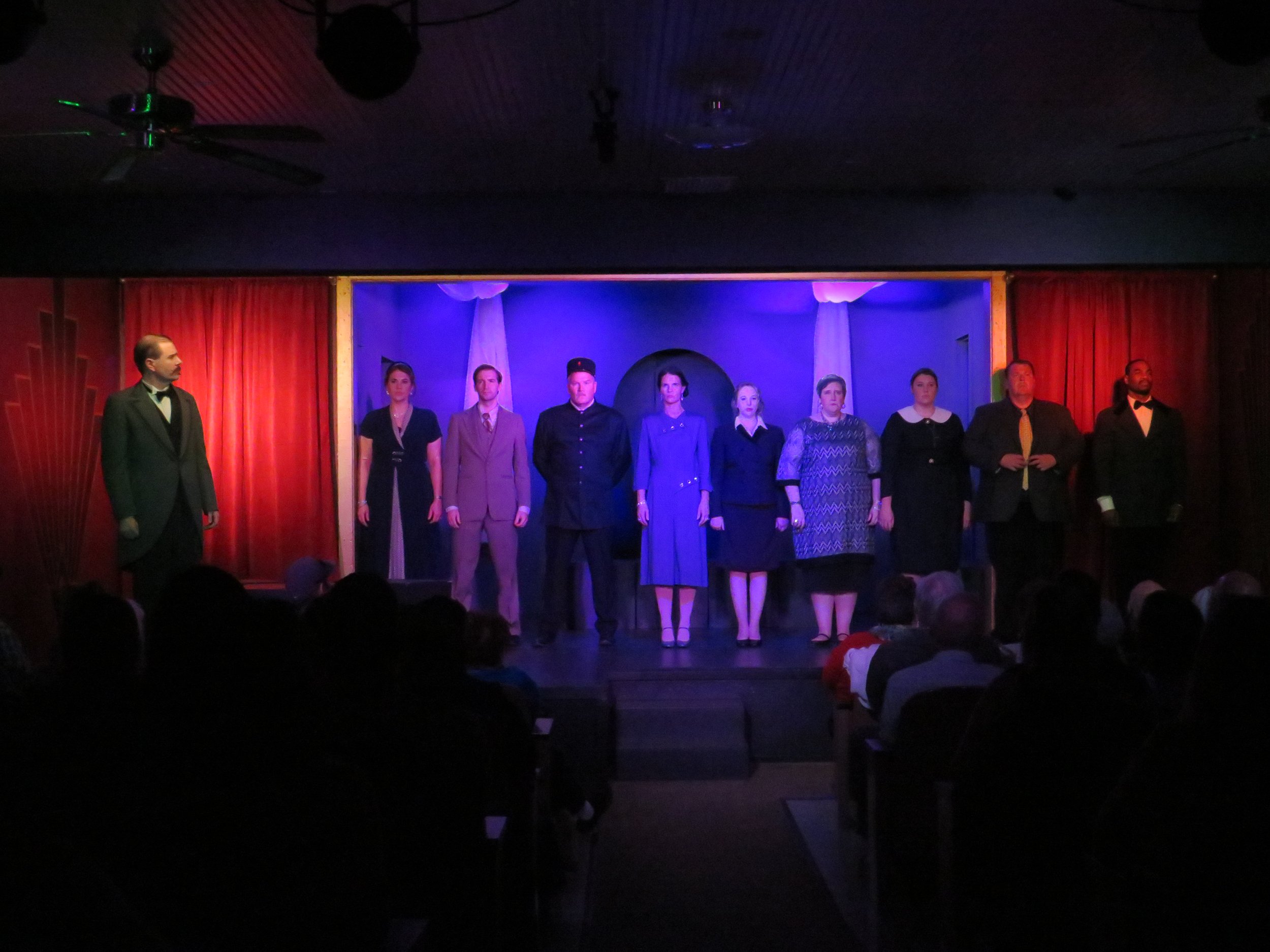 Actors standing on a stage, performing in a theatrical production, with audience in the foreground.