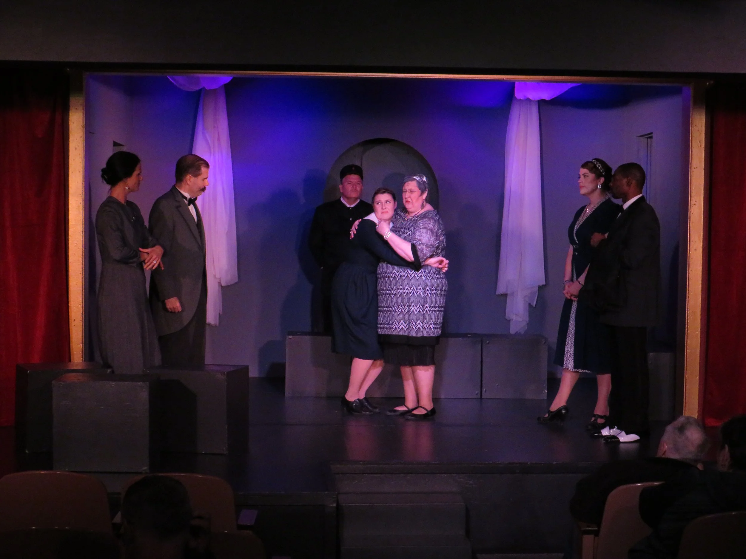 Theater scene with six actors on stage, three women and three men, engaged in a dramatic moment, with audience visible at the bottom.