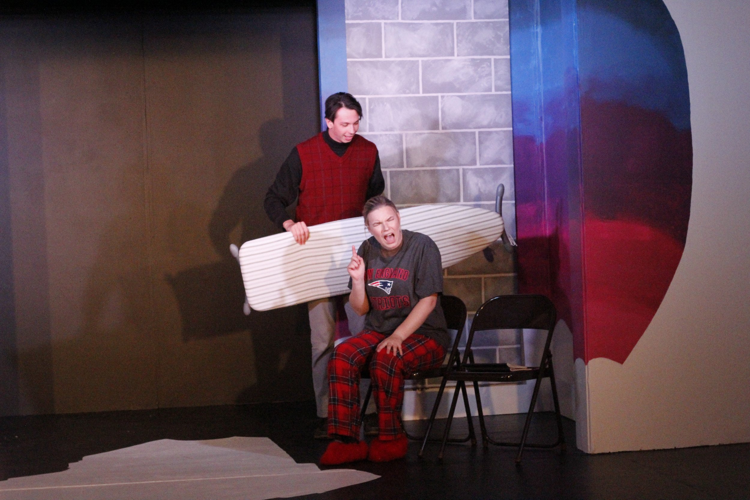 Two people performing on stage, one sitting on a chair dressed in pajamas with a New England Patriots T-shirt and plaid pants, making an expressive gesture with her mouth open, the other standing behind her holding a surfboard, in a set with a brick 