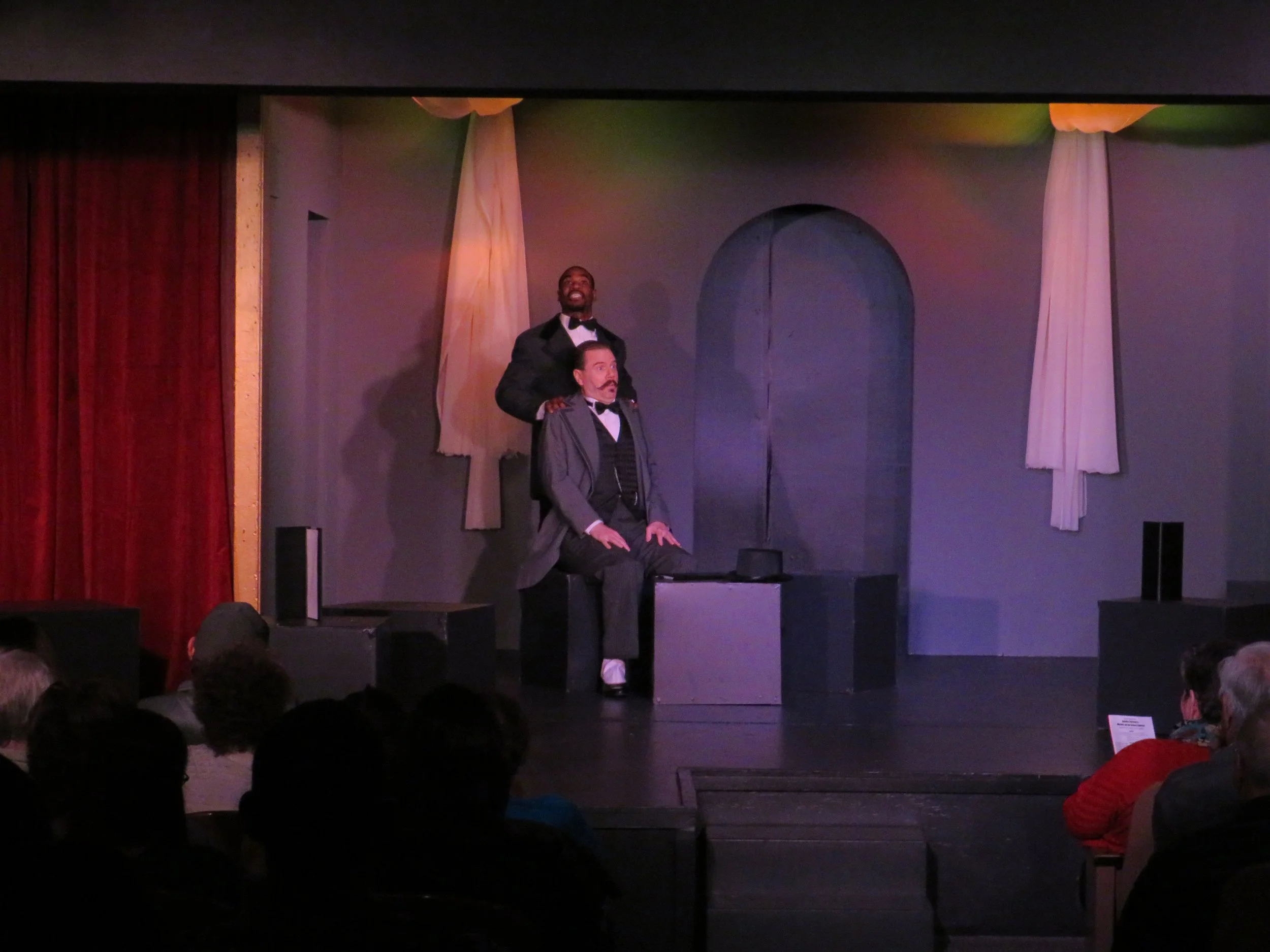The image shows two male actors on a theater stage, dressed in period costumes. One is standing behind the seated actor, placing his hands on his shoulders. The scene appears to be part of a theatrical performance with an audience watching.
