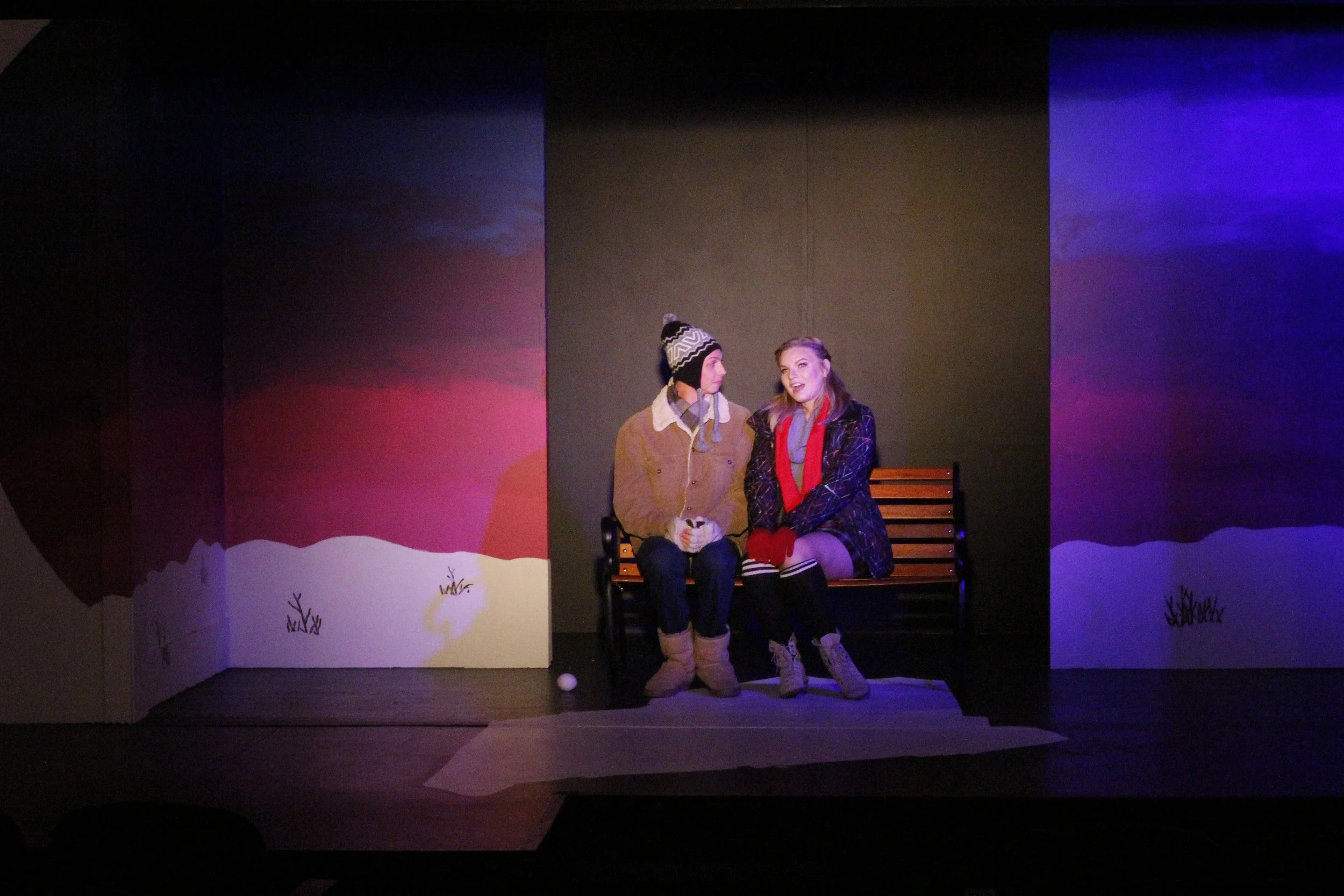 Two young actors sitting on a park bench during a theatrical performance, dressed in winter clothing, with a painted winter scene backdrop and colorful lighting.