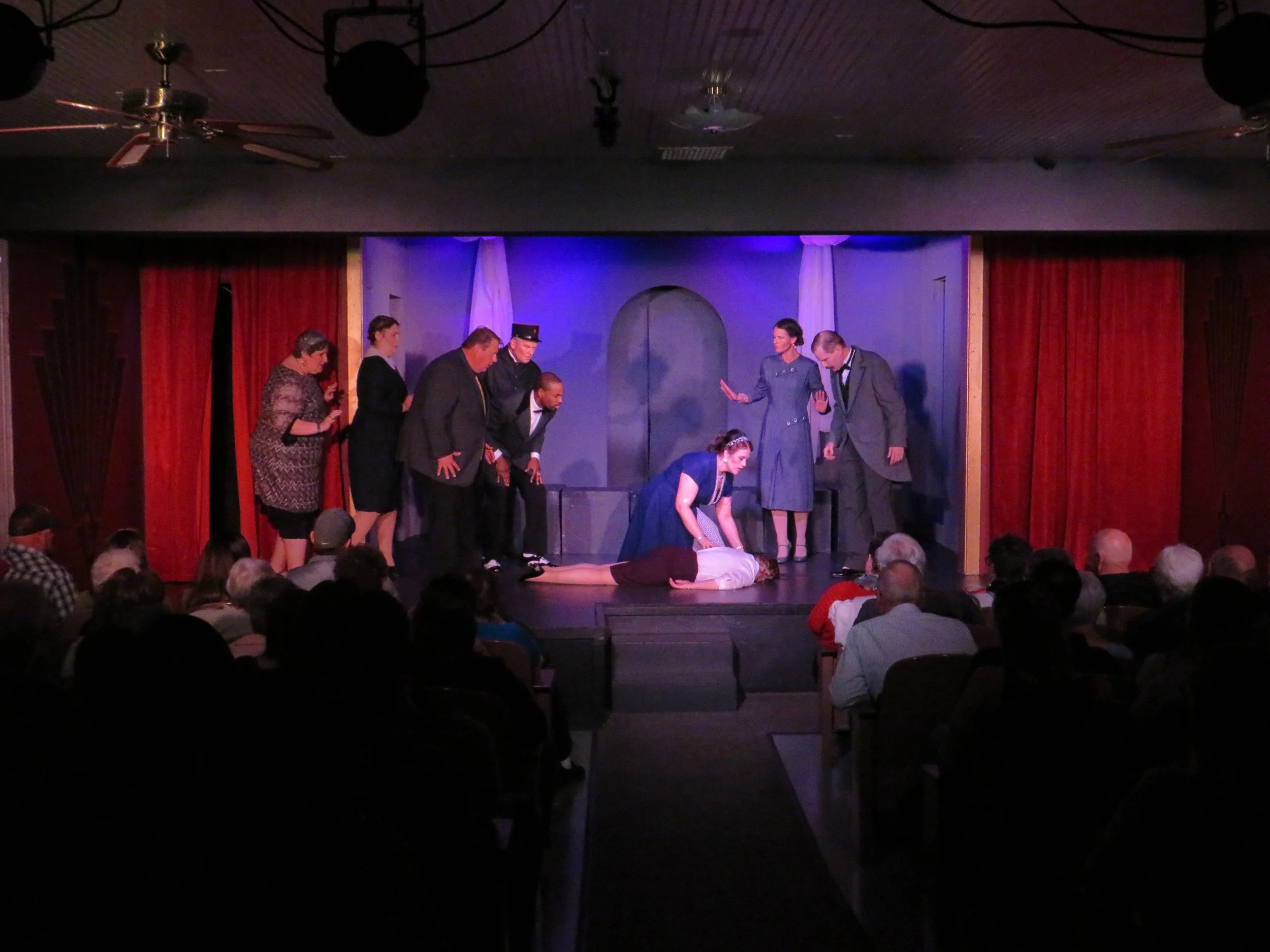 A stage play reenacting a scene with seven actors, including one lying on the floor and another kneeling beside her, with the audience watching from the darkened theater.