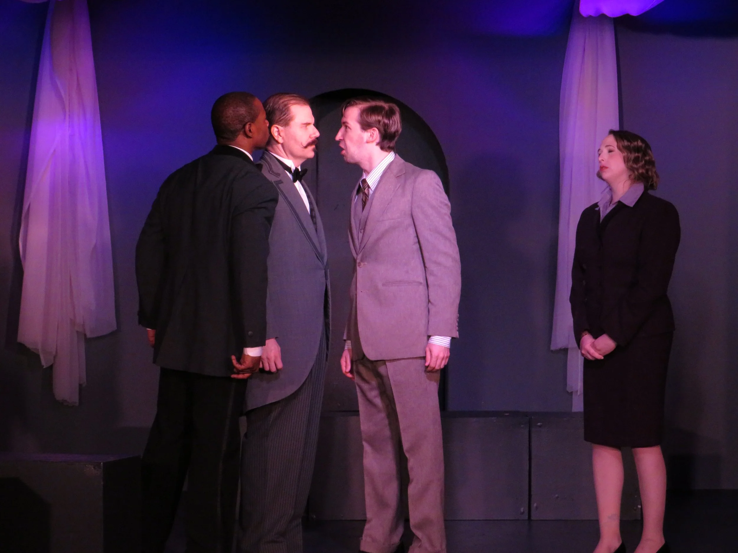 Four actors on stage in a theatrical scene, two men in suits facing each other closely, one man with a mustache and the other with short hair, a woman in a dark suit standing apart watching, and another man in a striped suit observing the confrontati