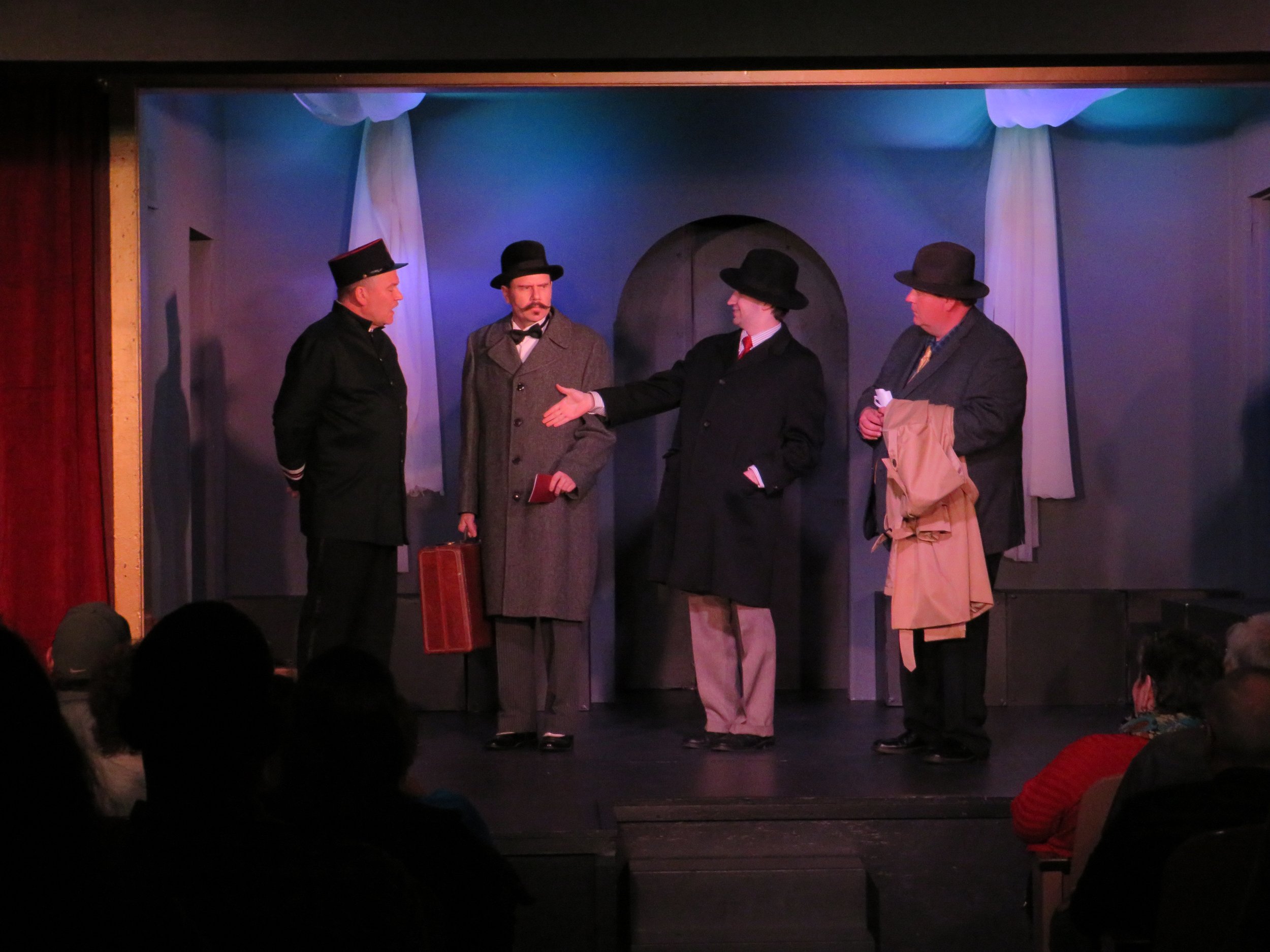 Four actors dressed in vintage clothing on stage, engaged in a scene, with audience members visible in the foreground.
