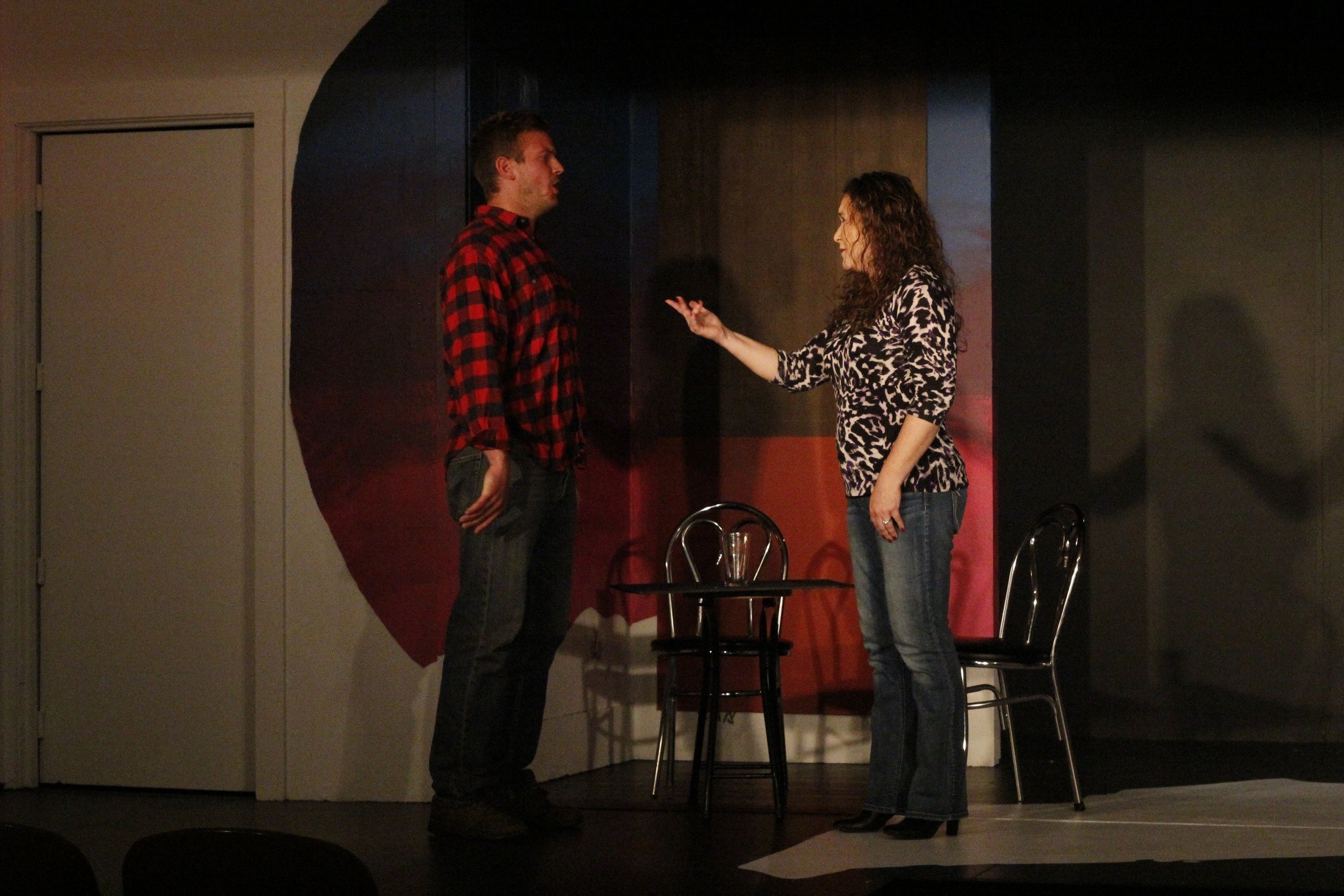 A man and woman are on stage engaging in a conversation. The man is wearing a red and black checkered shirt, standing with his hands in his pockets. The woman, wearing a leopard print blouse and jeans, is gesturing with her right hand. There are two 